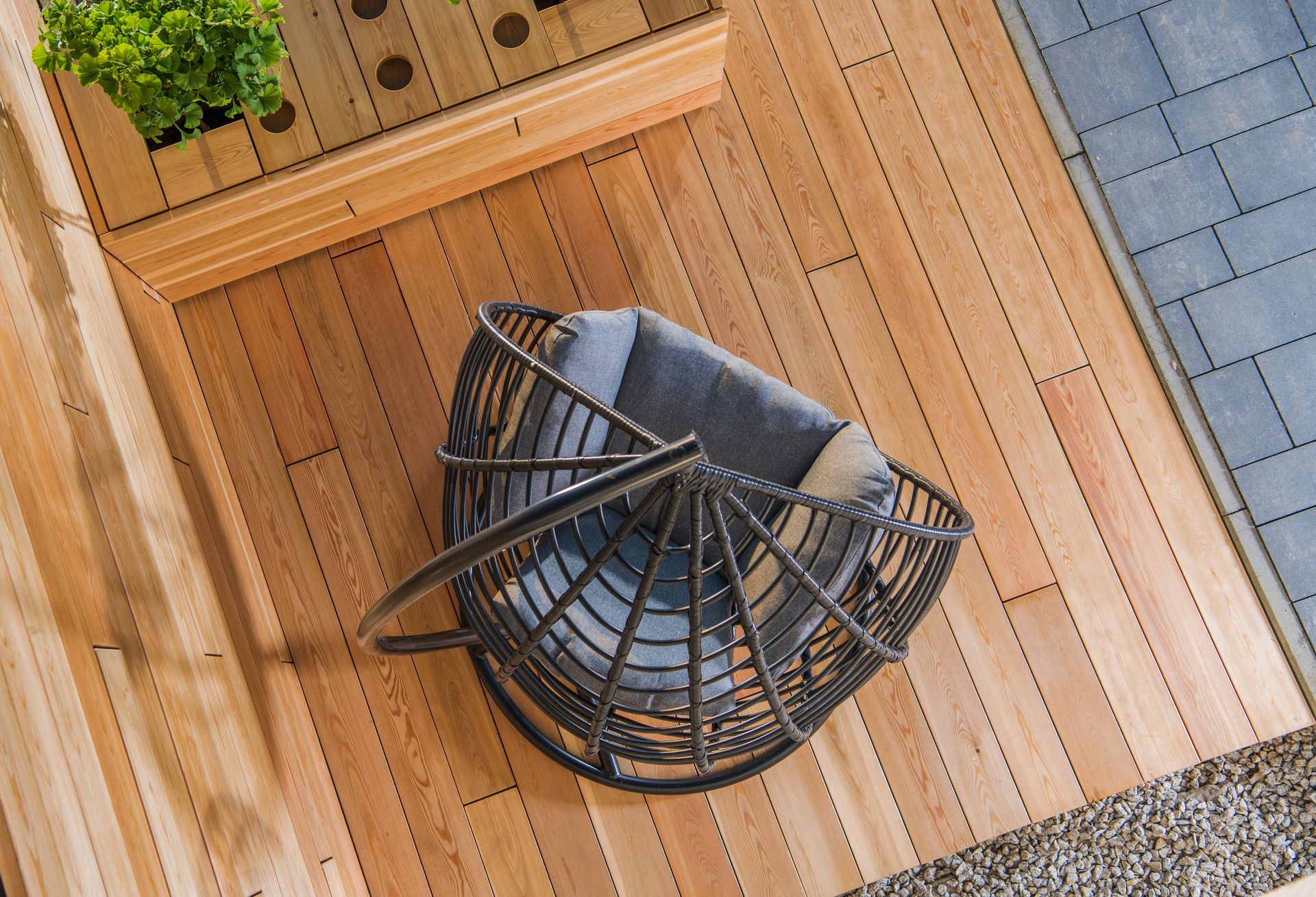 Overhead view of a wicker chair with cushions on a wooden deck, near a planter and gray pavers.