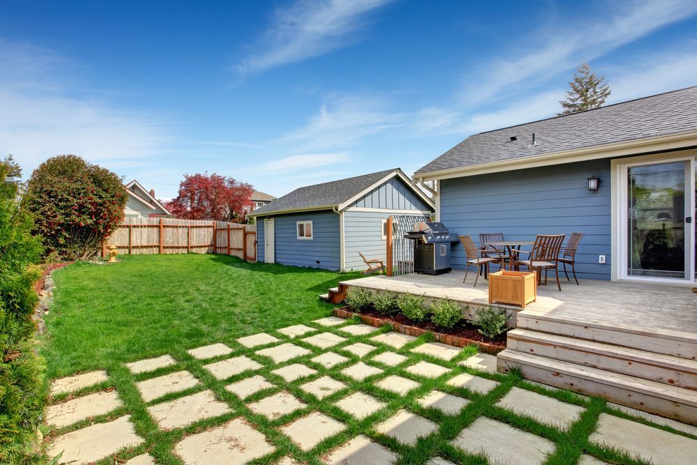 Backyard patio with stone path, green lawn, and blue house under a clear sky
