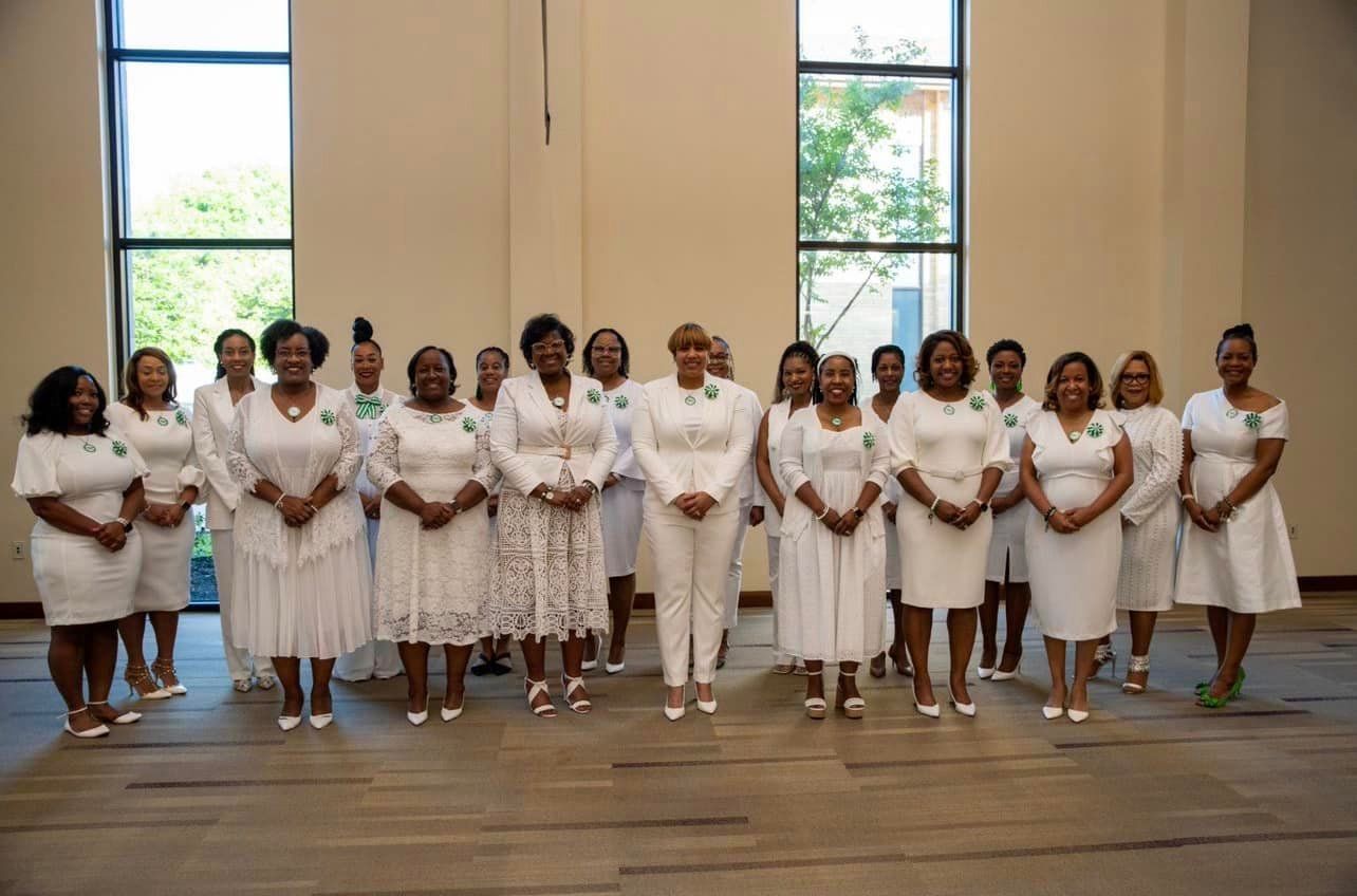 Group of women in white attire, posing indoors near windows. Each wears a green pin.