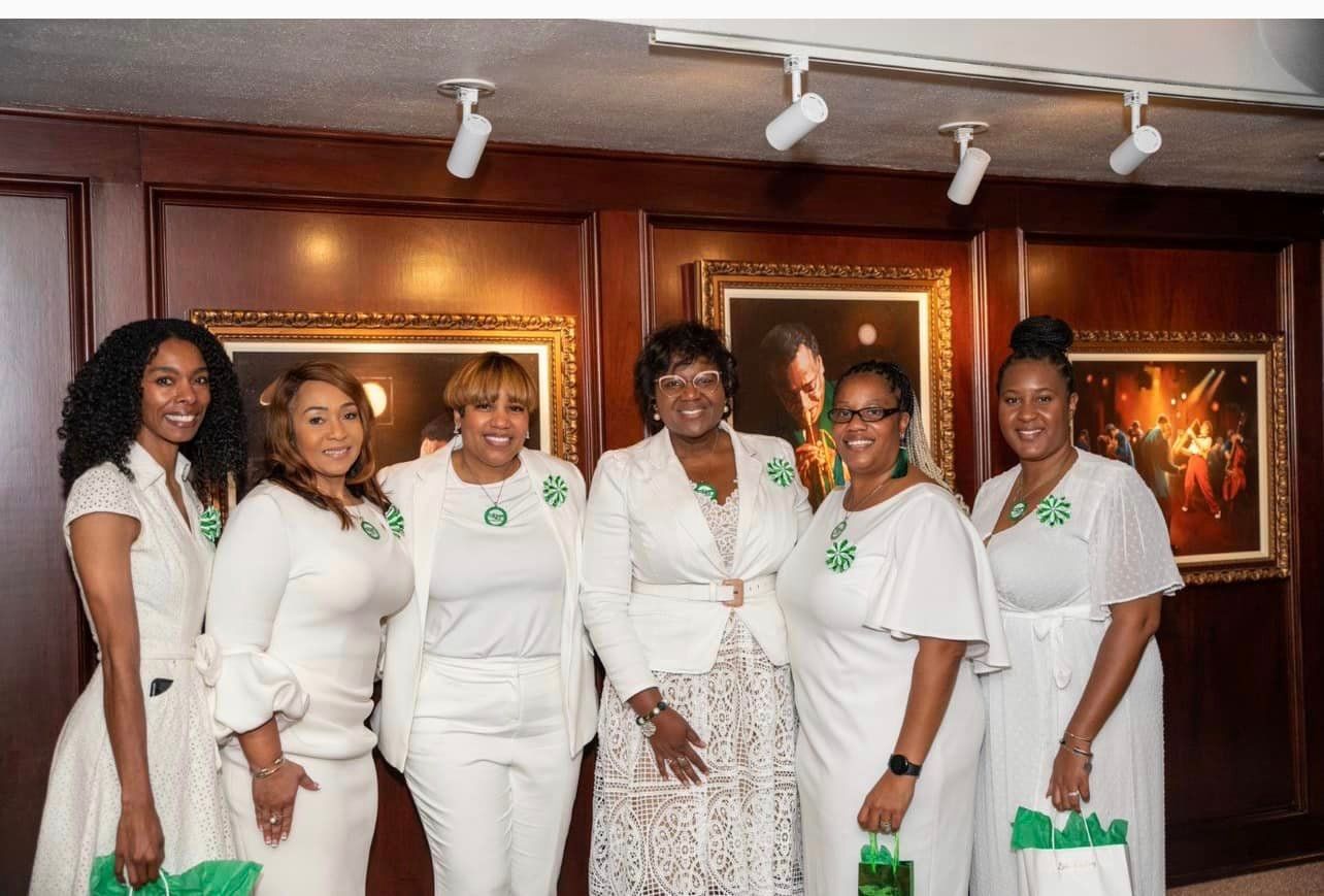 Six women in white attire pose in front of artwork. All wear green accents, smile, and hold green bags.