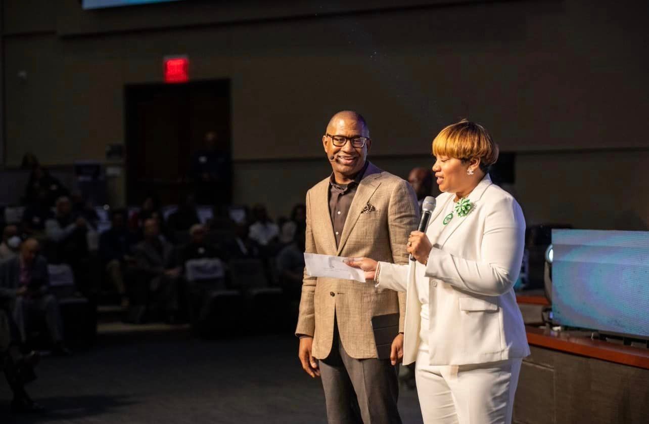 Man and woman on stage, woman holding a microphone. Auditorium with audience visible.