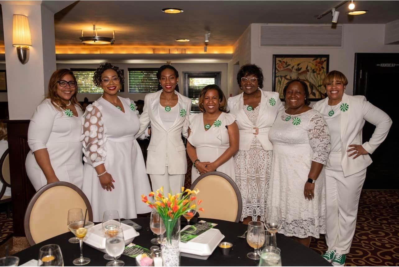 Group of women in white outfits, green accents, standing near a dining table in a restaurant.