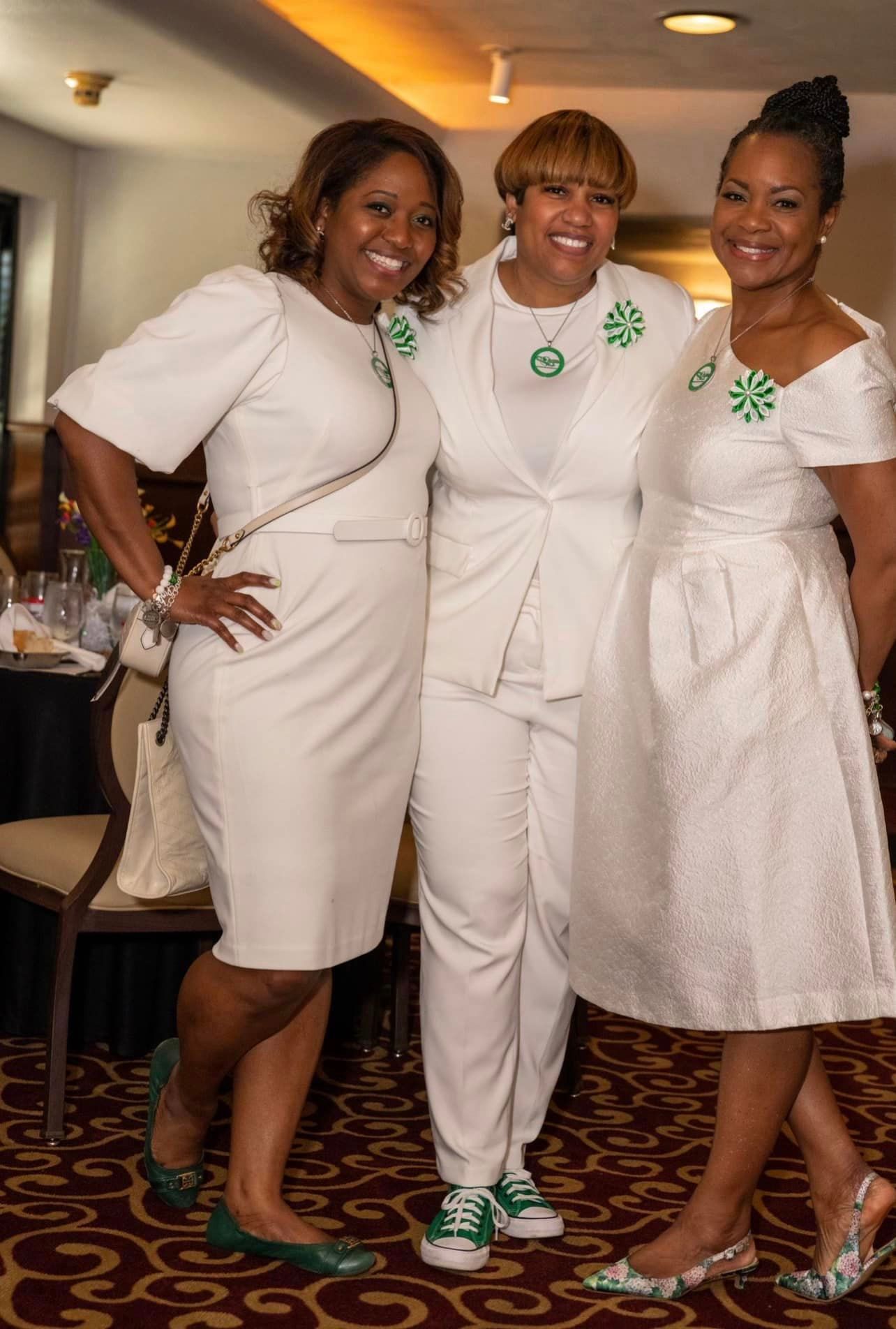 Three women in white outfits pose together. They are wearing green accents and smiling, indoors at an event.