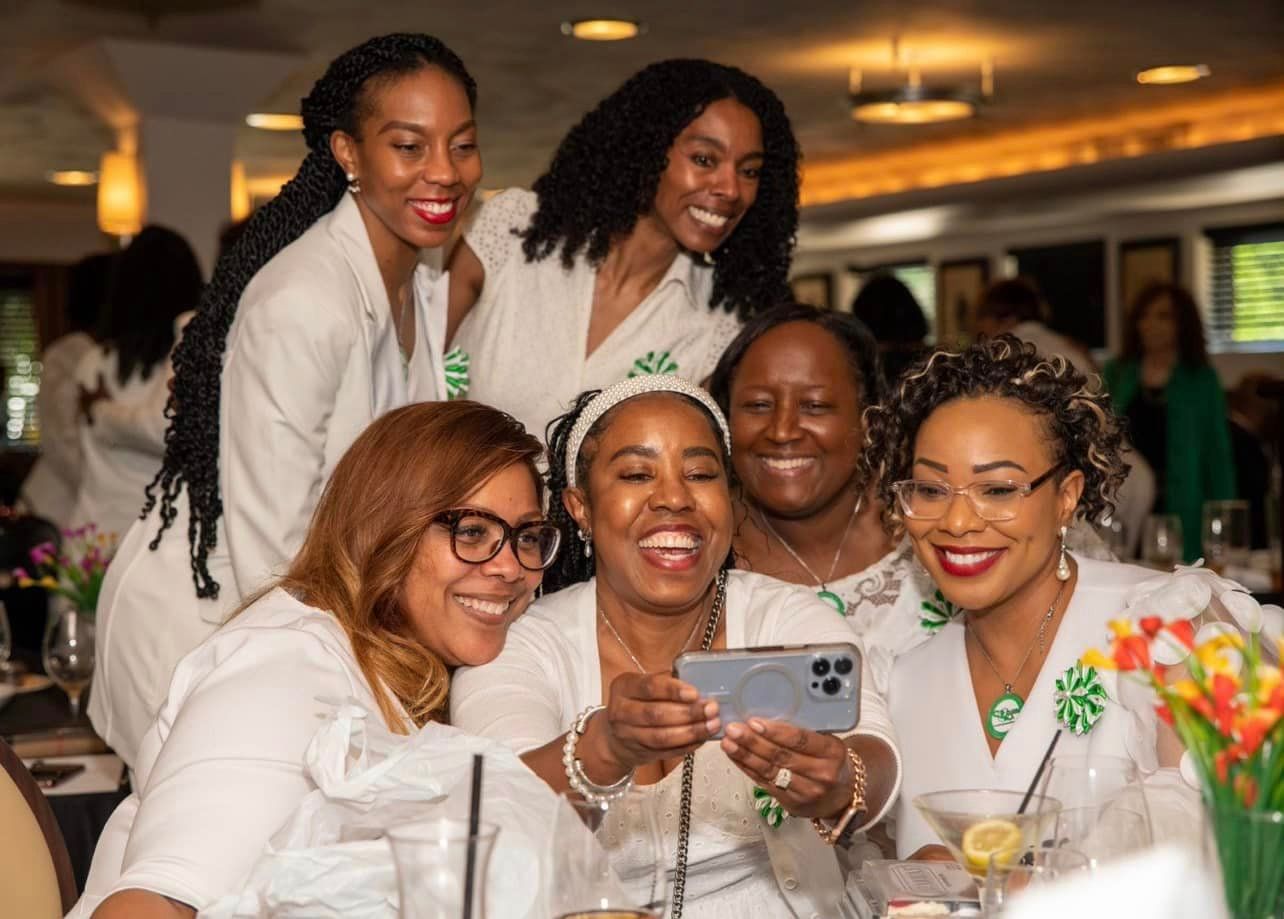 Group of women smiling, taking a selfie at a restaurant. All wearing white and green outfits, some with flowers.