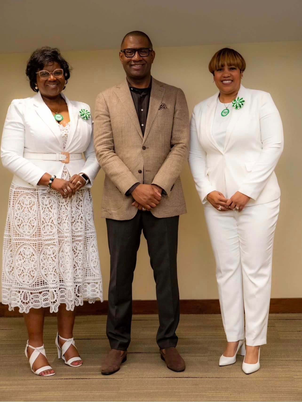 Three people pose for a photo. A man in a brown suit stands between two women in white suits.