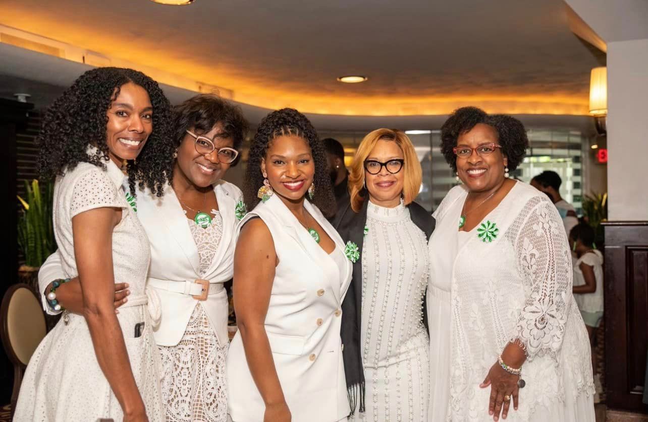 Five women in white outfits, smiling, posing together indoors with greenery in the background.
