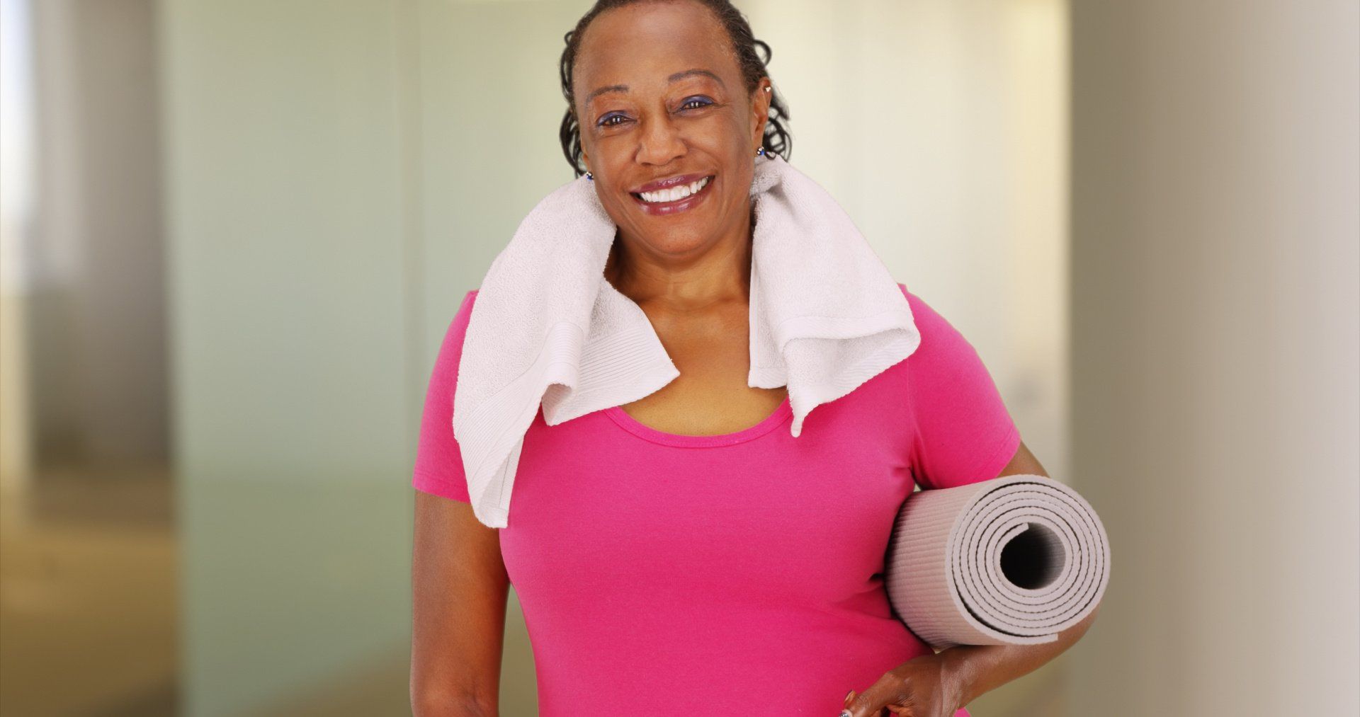 Woman holding yoga mat, towel around neck, smiling.