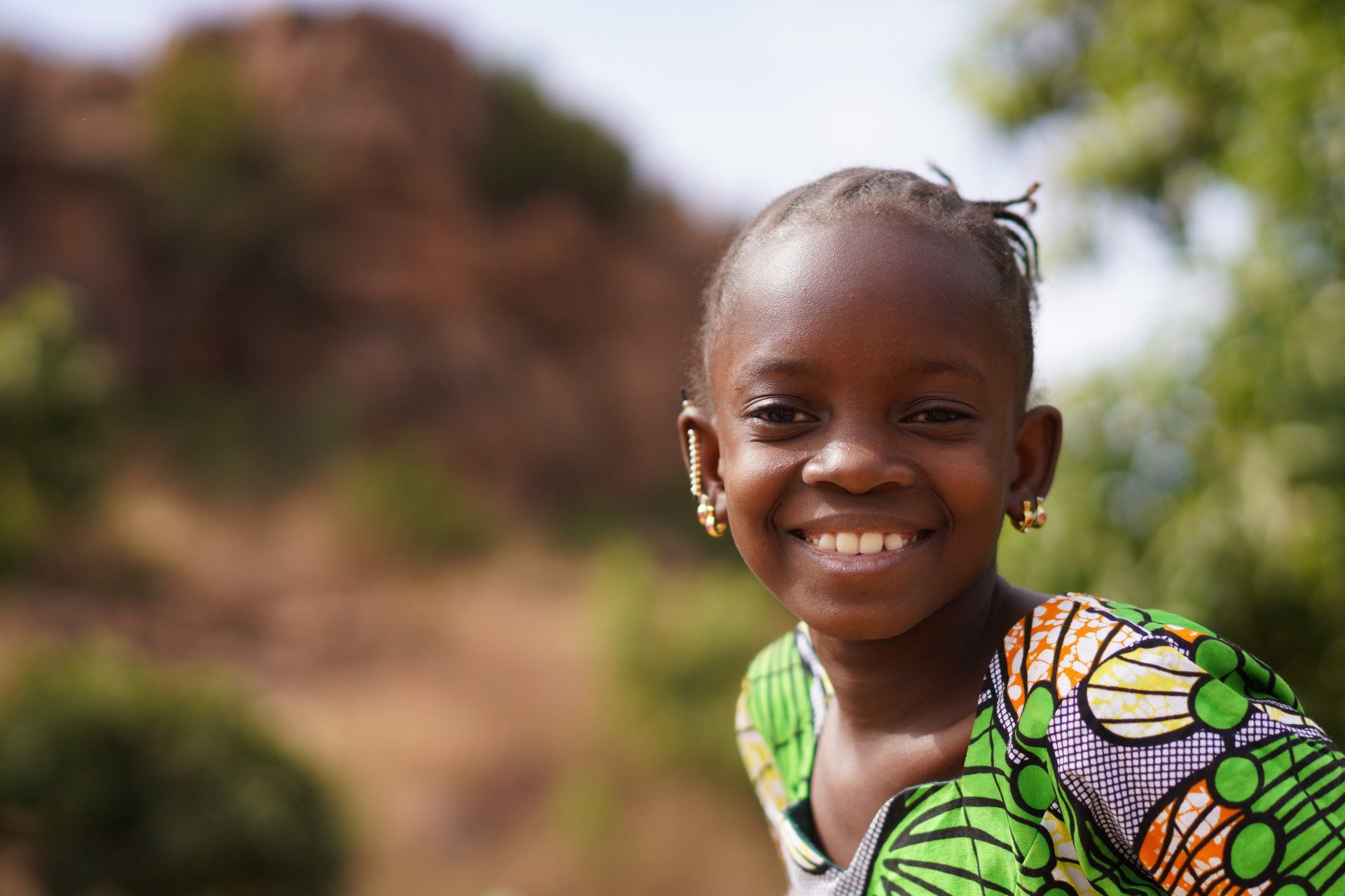 Girl smiling, wearing patterned green top, gold earrings, outdoors with blurry background.