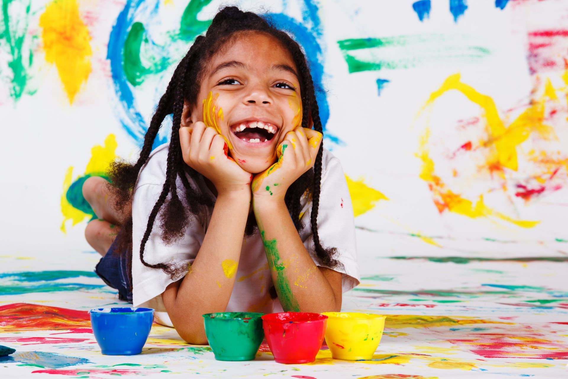 Girl smiling, covered in paint, lying on floor with colorful paint splatters and bowls.
