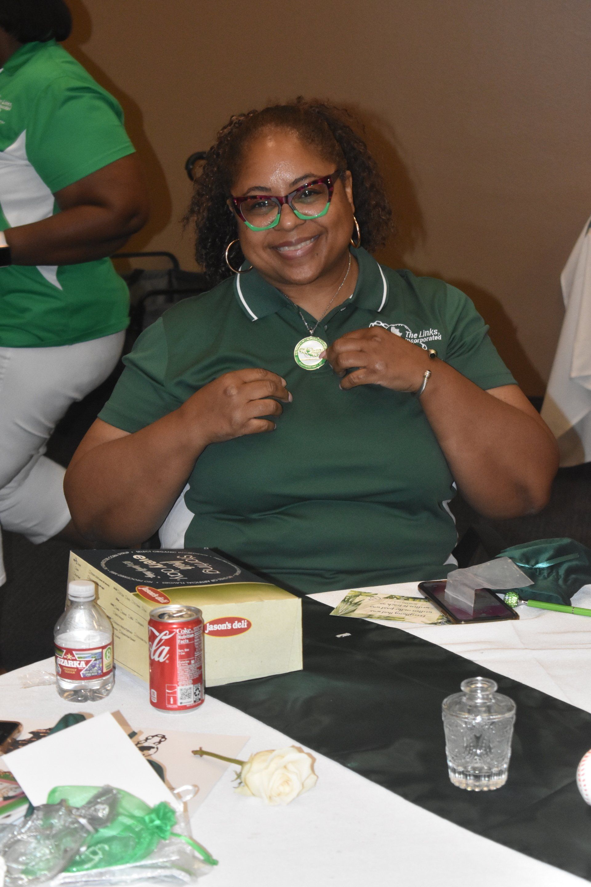 Woman wearing a green polo shirt and glasses smiles while adjusting a pin. Sitting at a table.