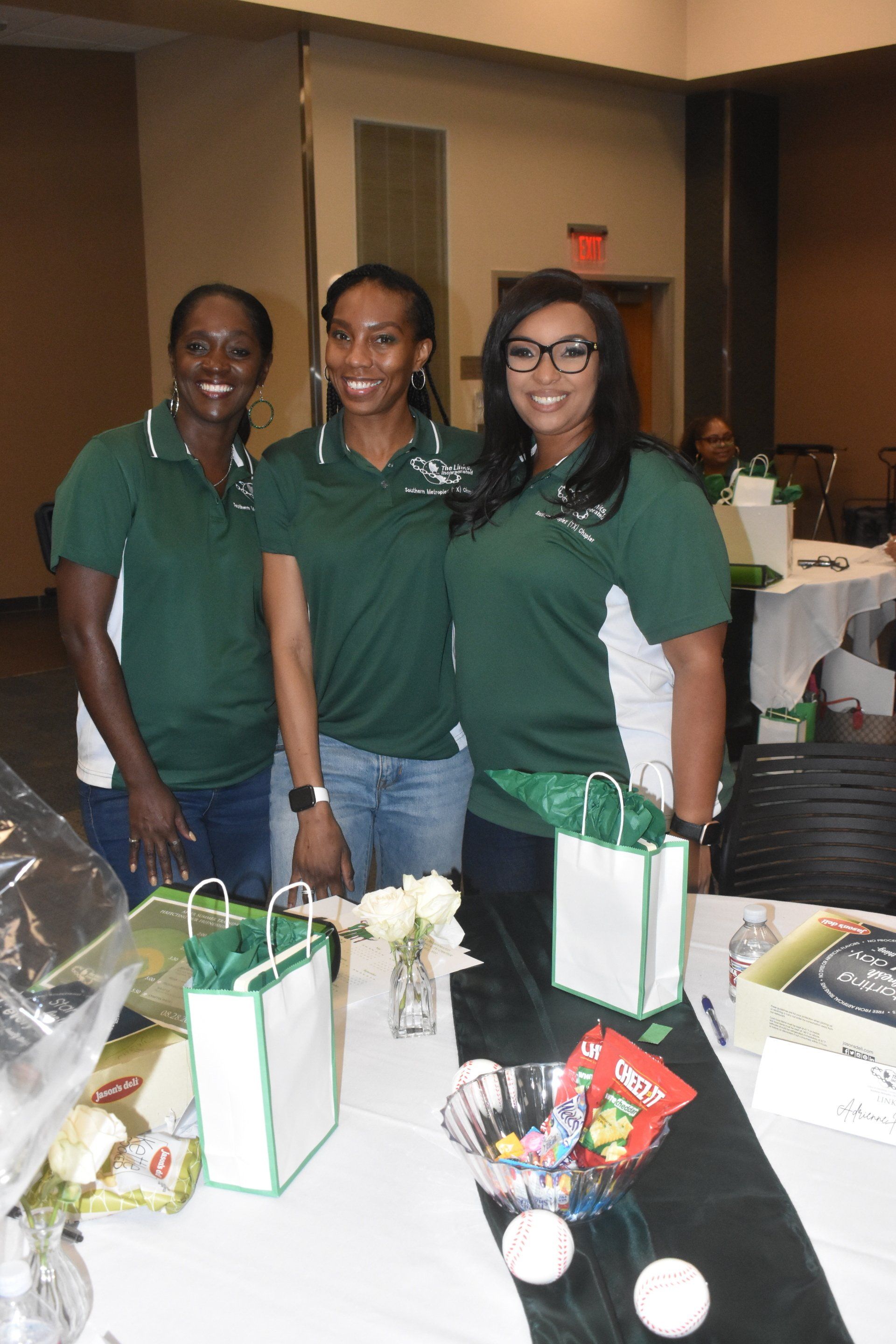 Three women smiling at a table, wearing green shirts. Event setting with gifts.