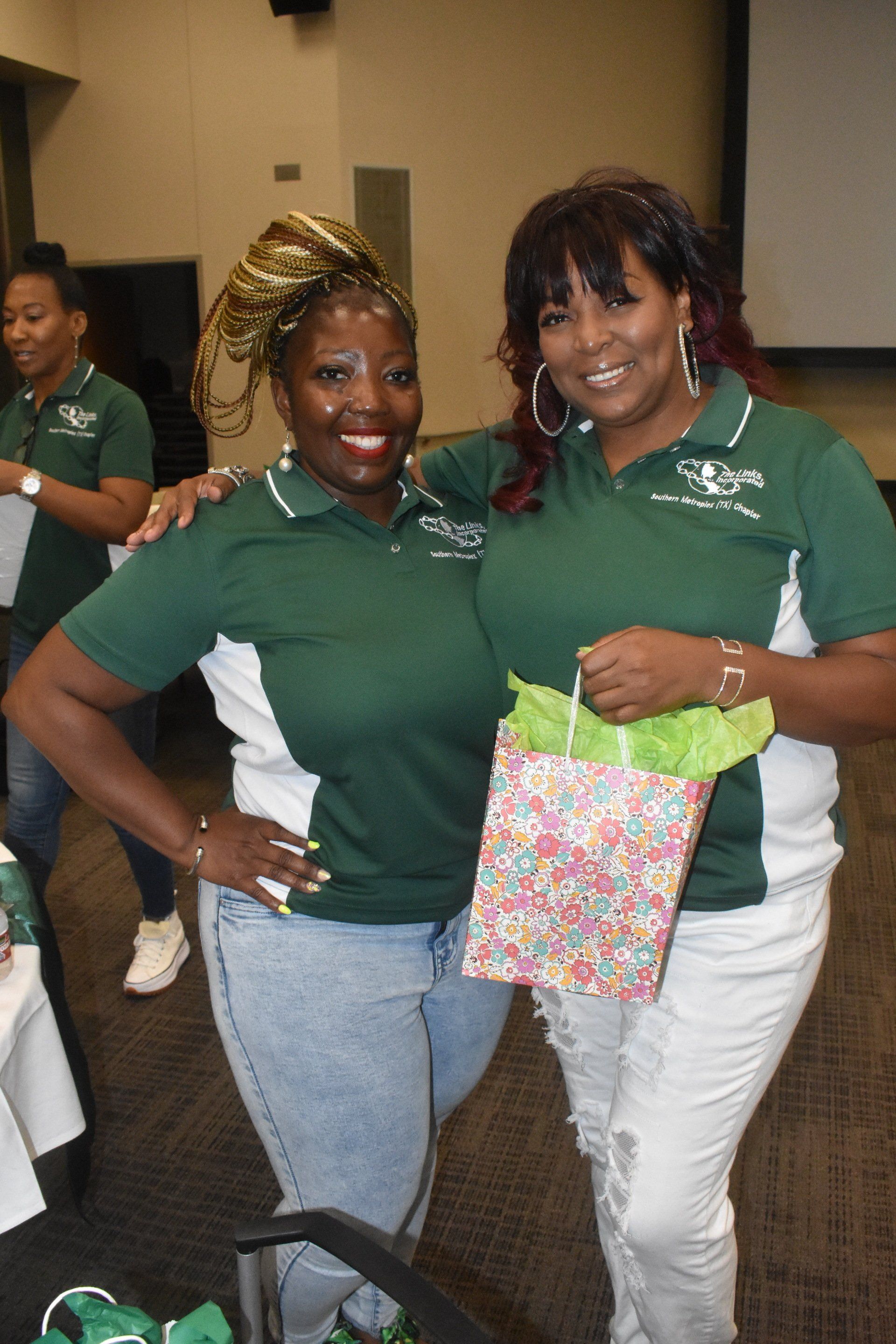 Two women smiling, posing for a photo. One holds a gift bag. Both wearing green shirts with logos.