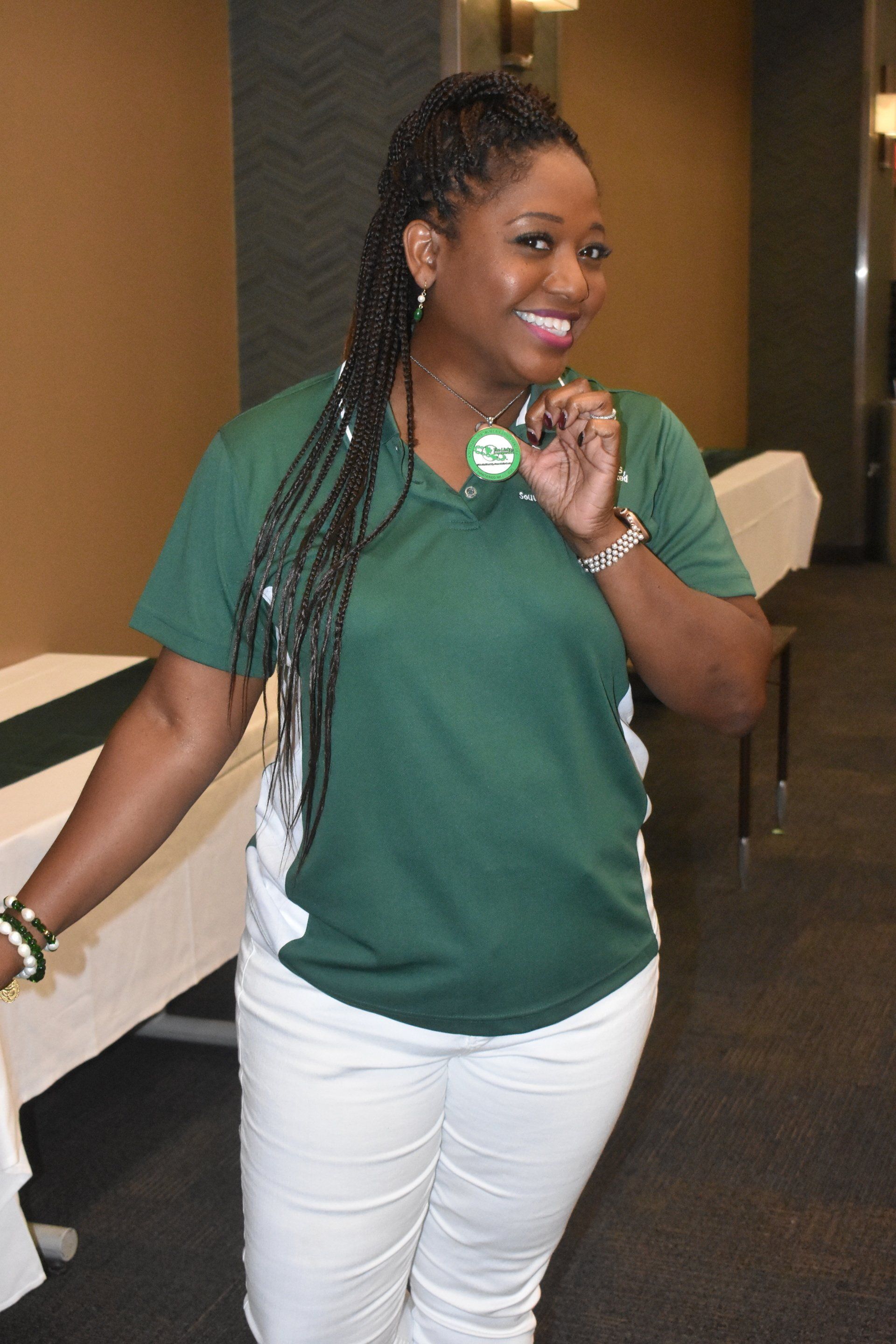 Woman in green shirt and white pants, smiling, holding a button, standing by a table.