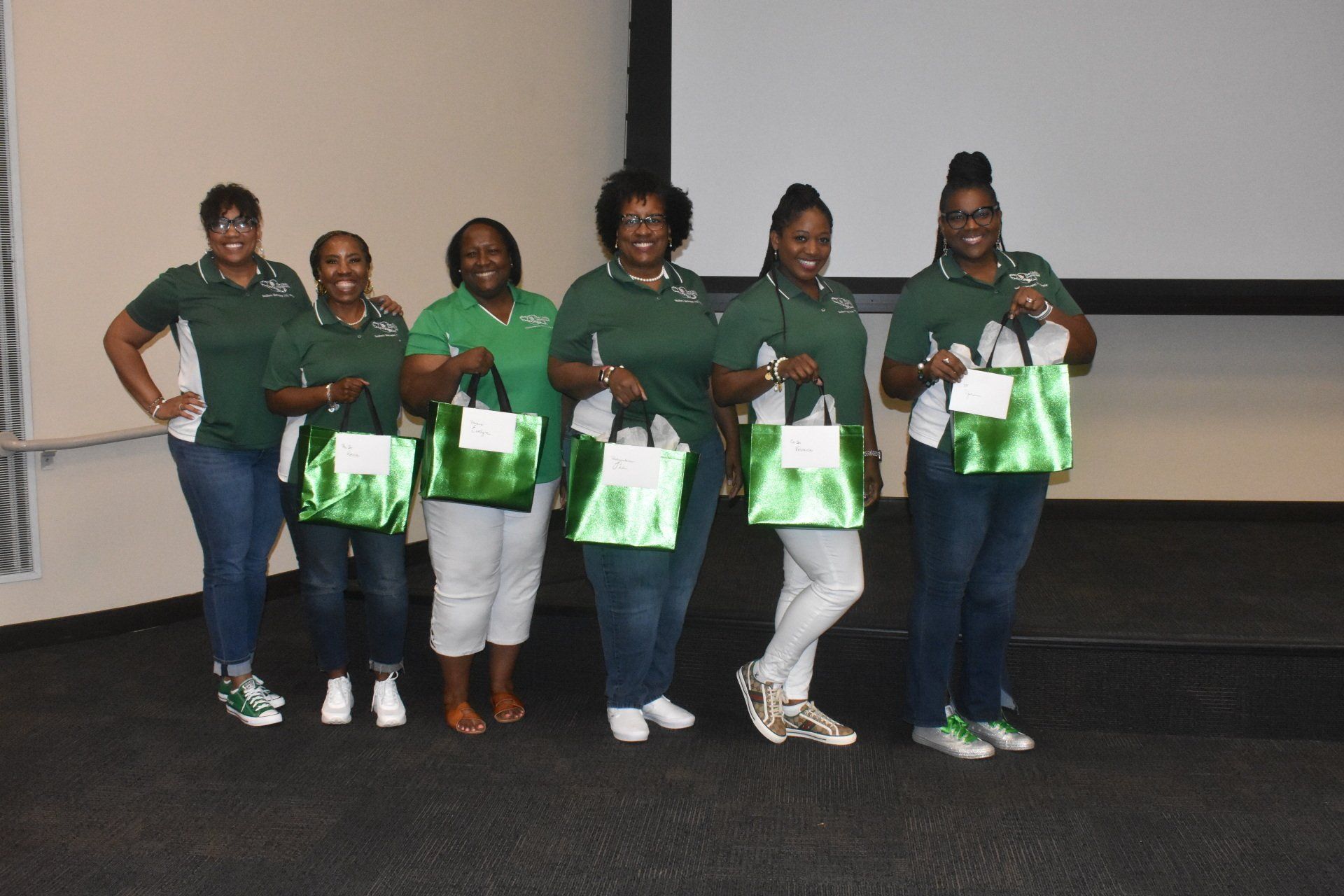 Six people stand holding green bags in front of a screen, wearing green and white shirts.