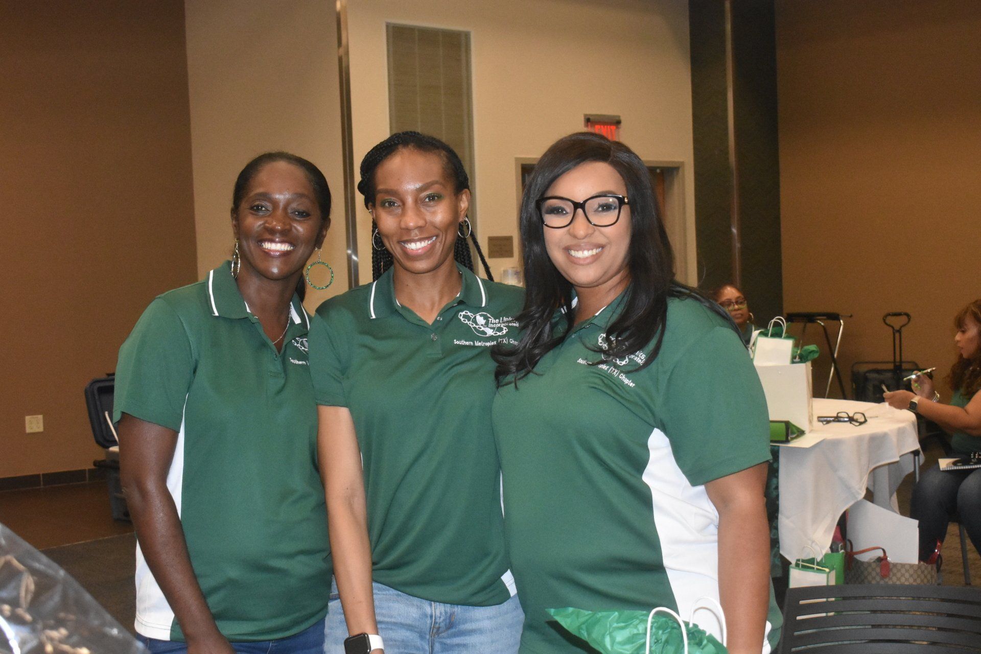 Three people in green shirts smile. They are indoors, possibly at an event.