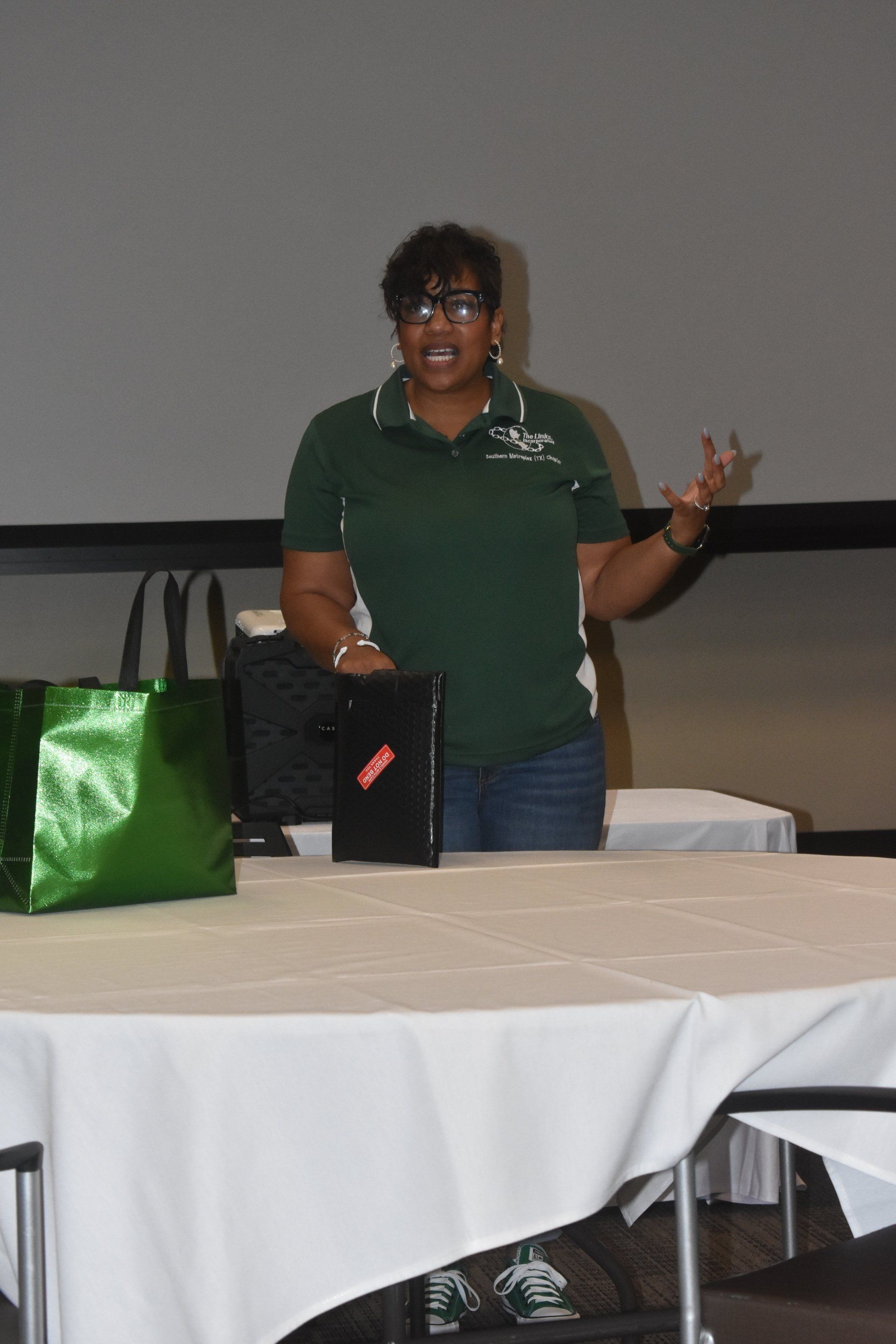 Person speaking at a table, wearing a green polo shirt and jeans, gesturing with hands. A laptop and bags sit on the table.