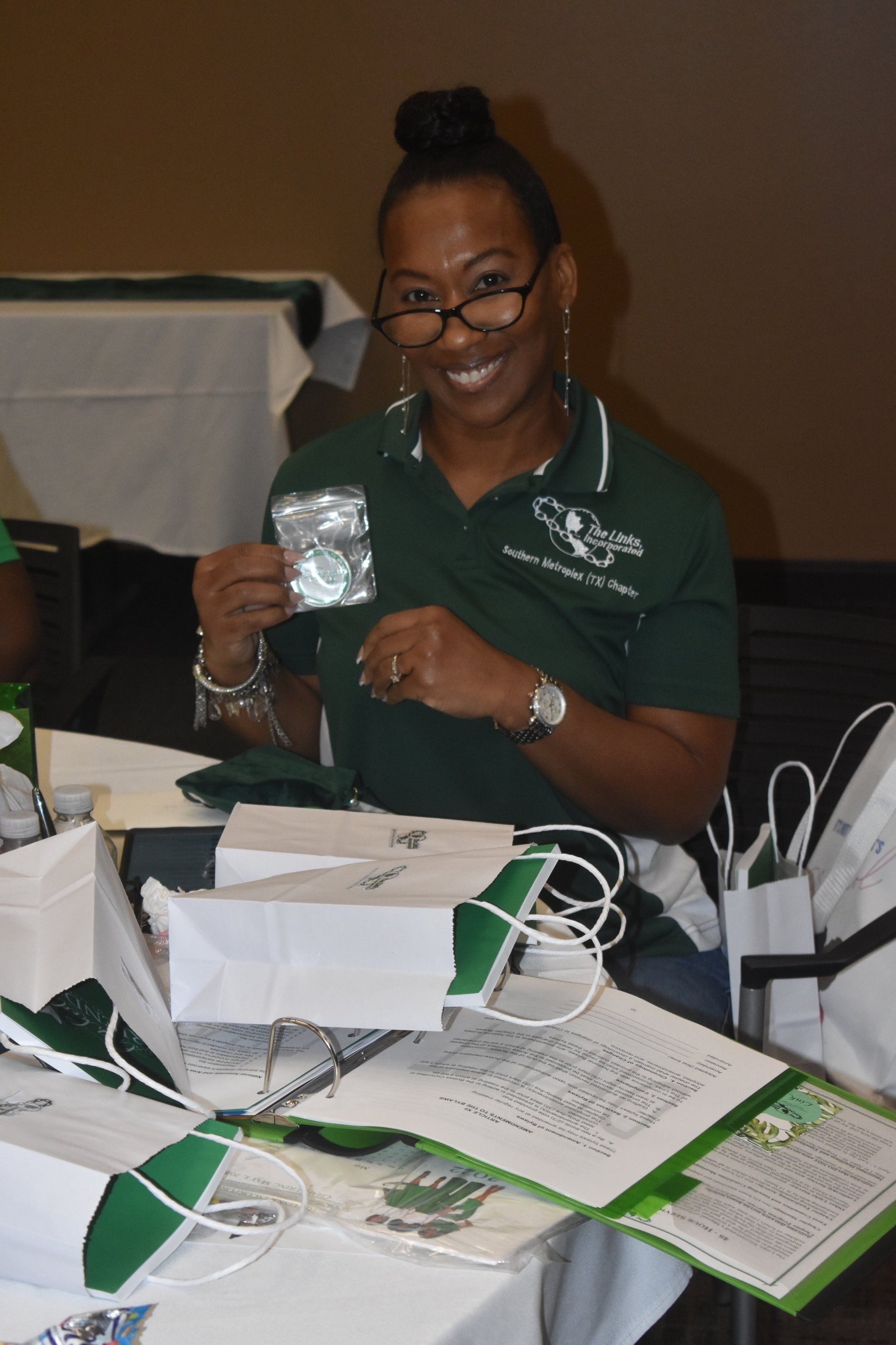 Woman in green polo holds small, foil-wrapped item. She smiles, sitting at a table with white gift bags and papers.