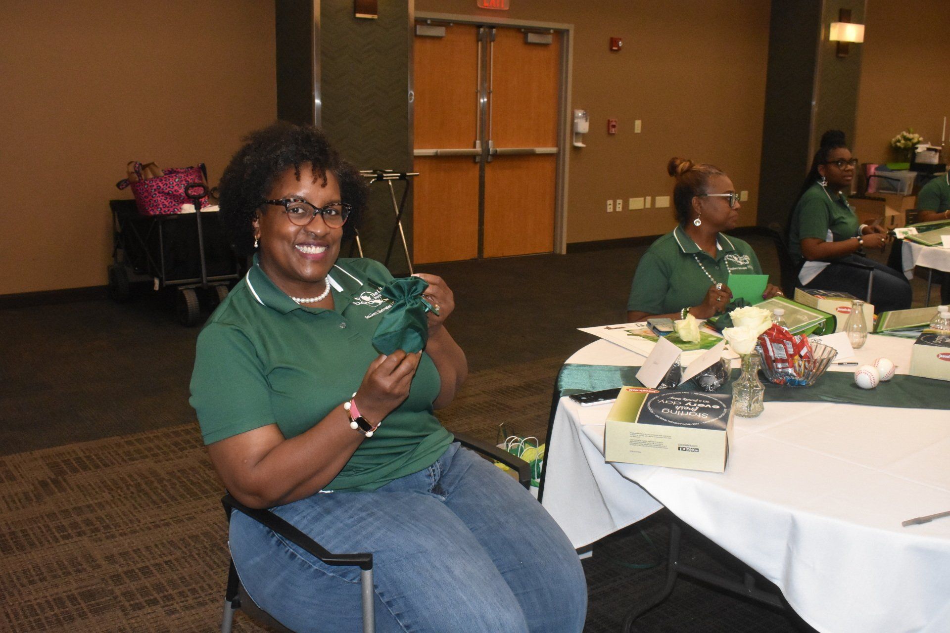 Woman in green shirt smiles, holding a green item, seated at a table with others in a conference room.