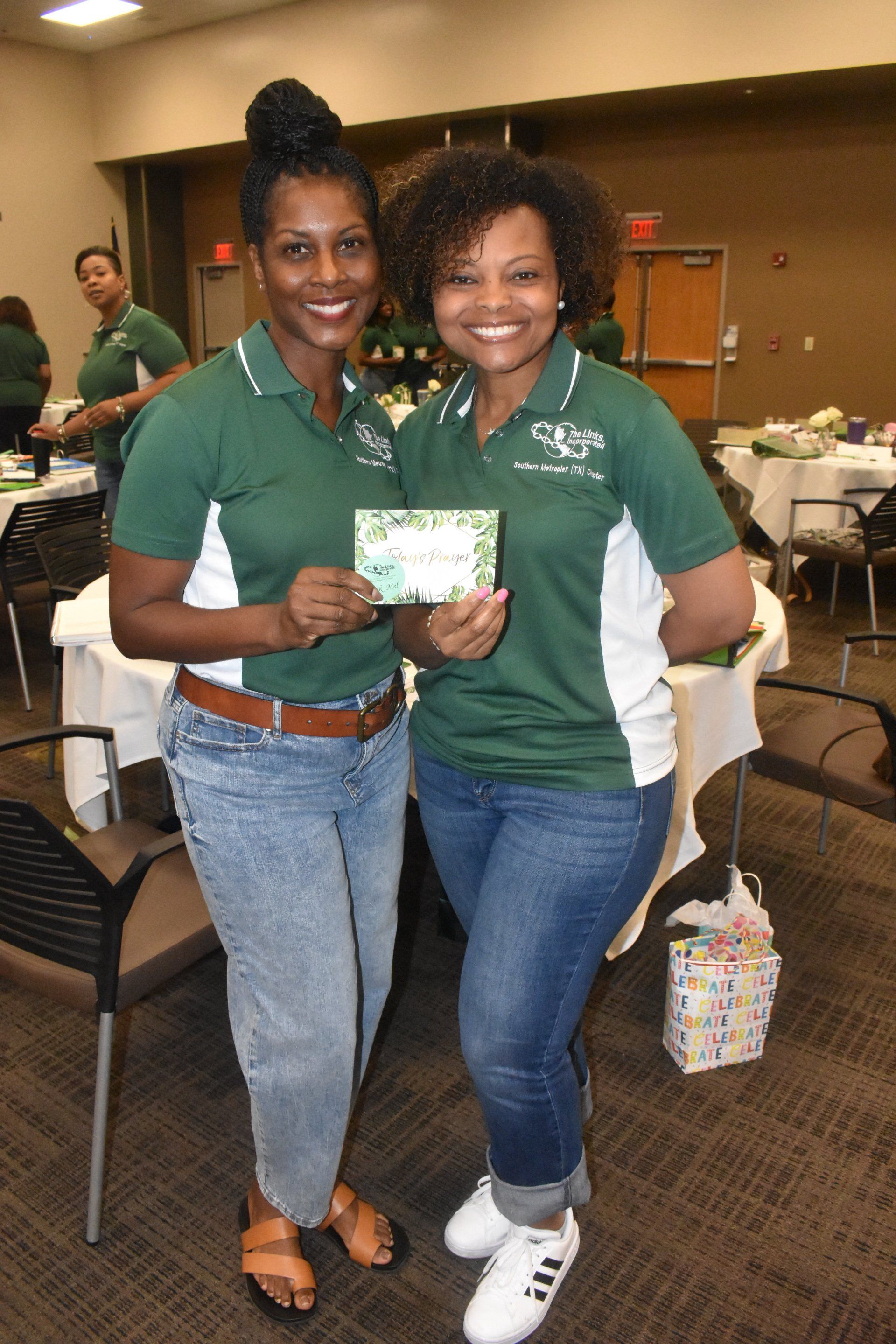 Two women in matching green shirts, holding a card, smiling in a room with tables and chairs.