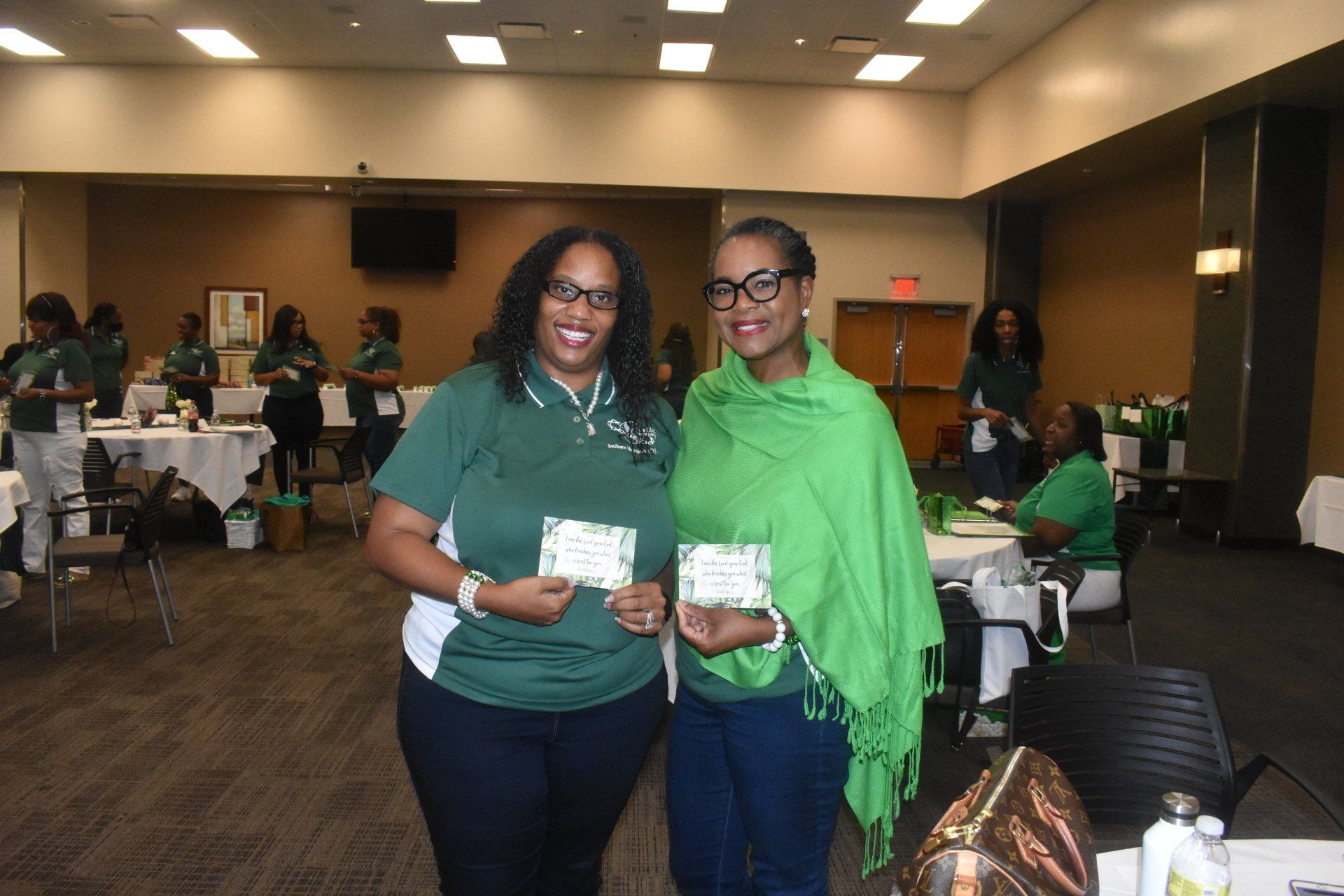 Two women in green outfits holding cards, smiling, in a room with tables and other people.
