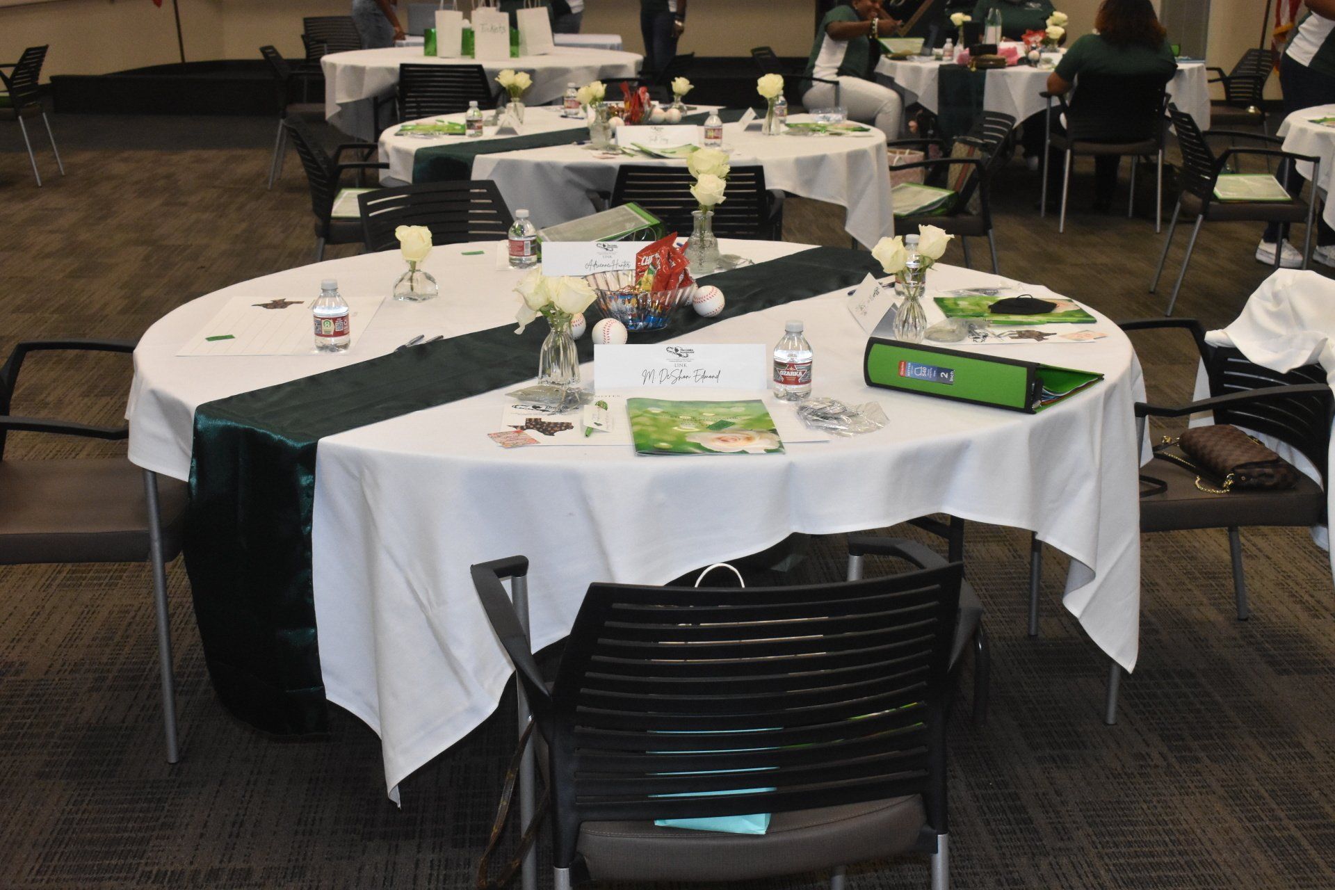 Tables set for an event, white tablecloths with green runners, vases of white flowers, bottled water, and event materials.