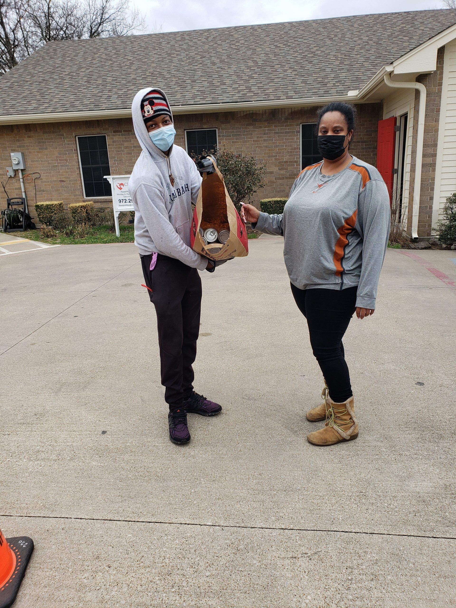 A person in a gray hoodie hands a brown paper bag to another person, outside a building with red doors.