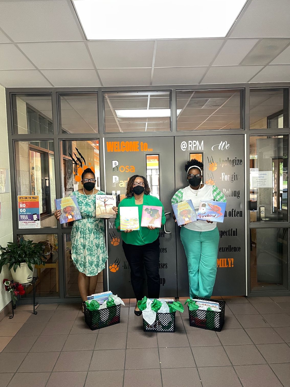 Three people holding books, standing in front of a school entrance, with baskets of books.