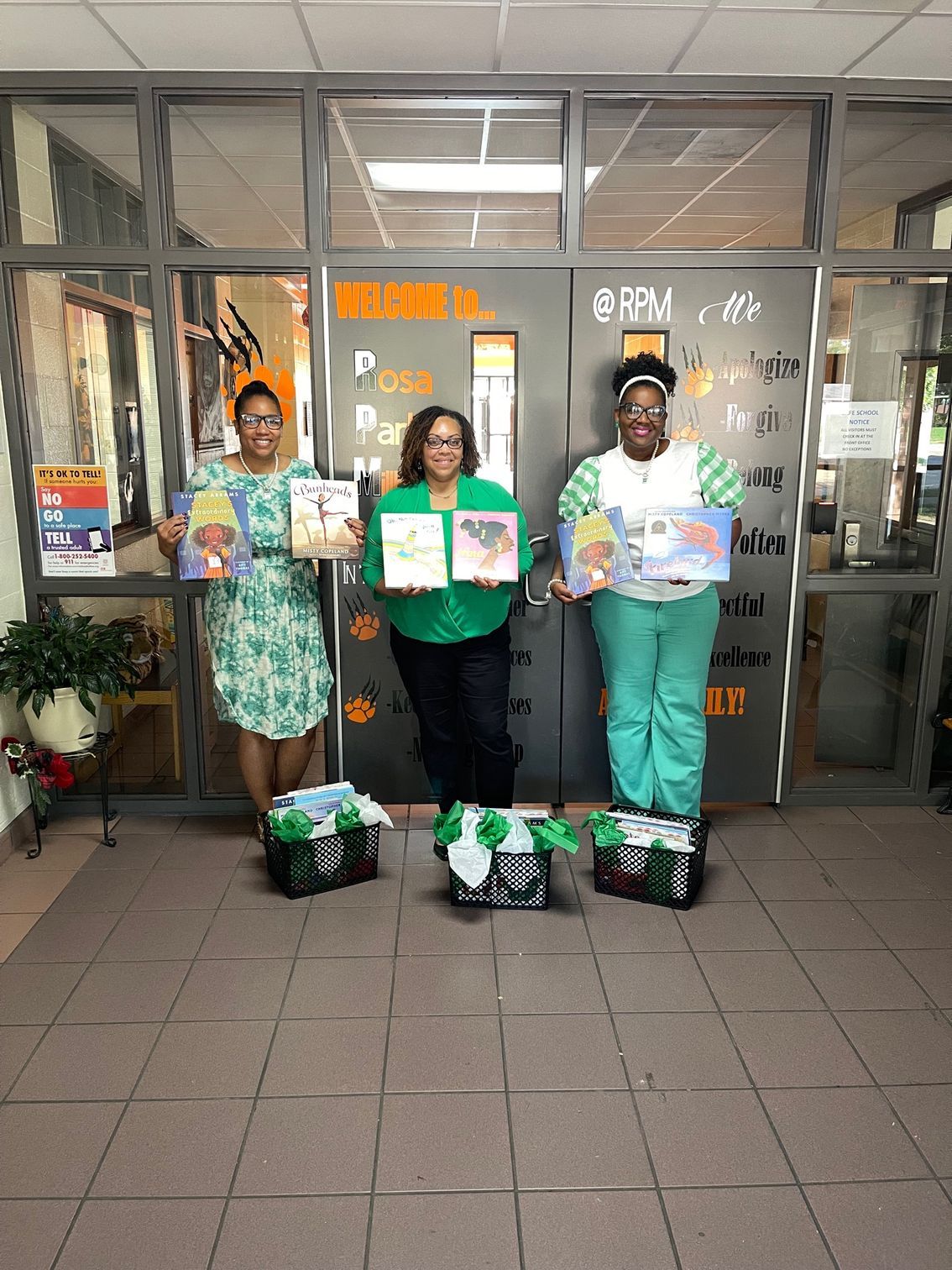 Three people holding books, standing near glass doors, with gift baskets in front.