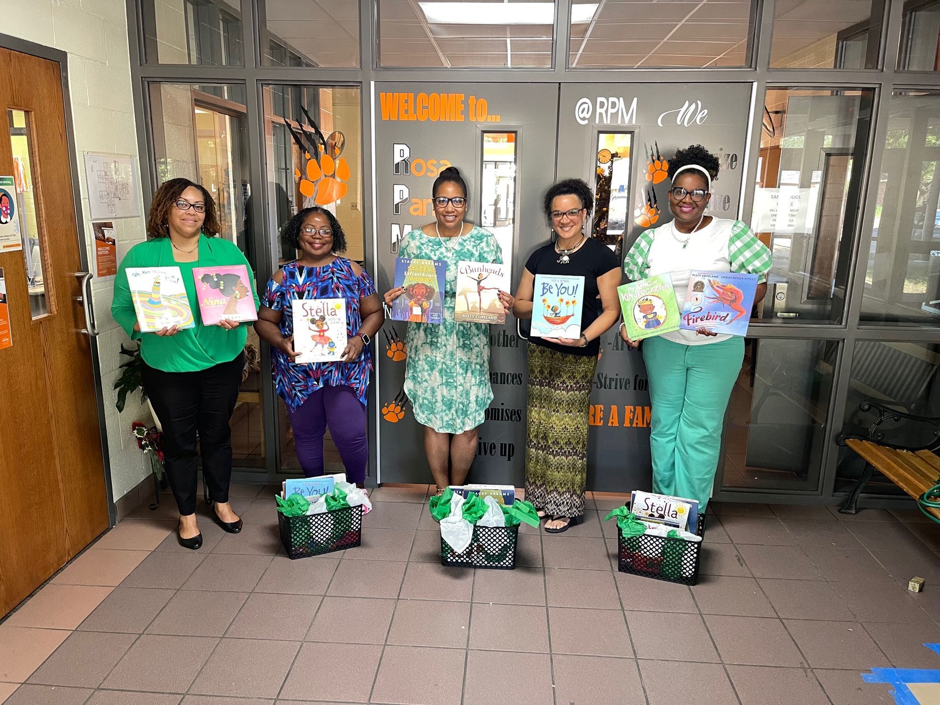 Five people hold books and stand near baskets. They are in a school hallway near a door with 