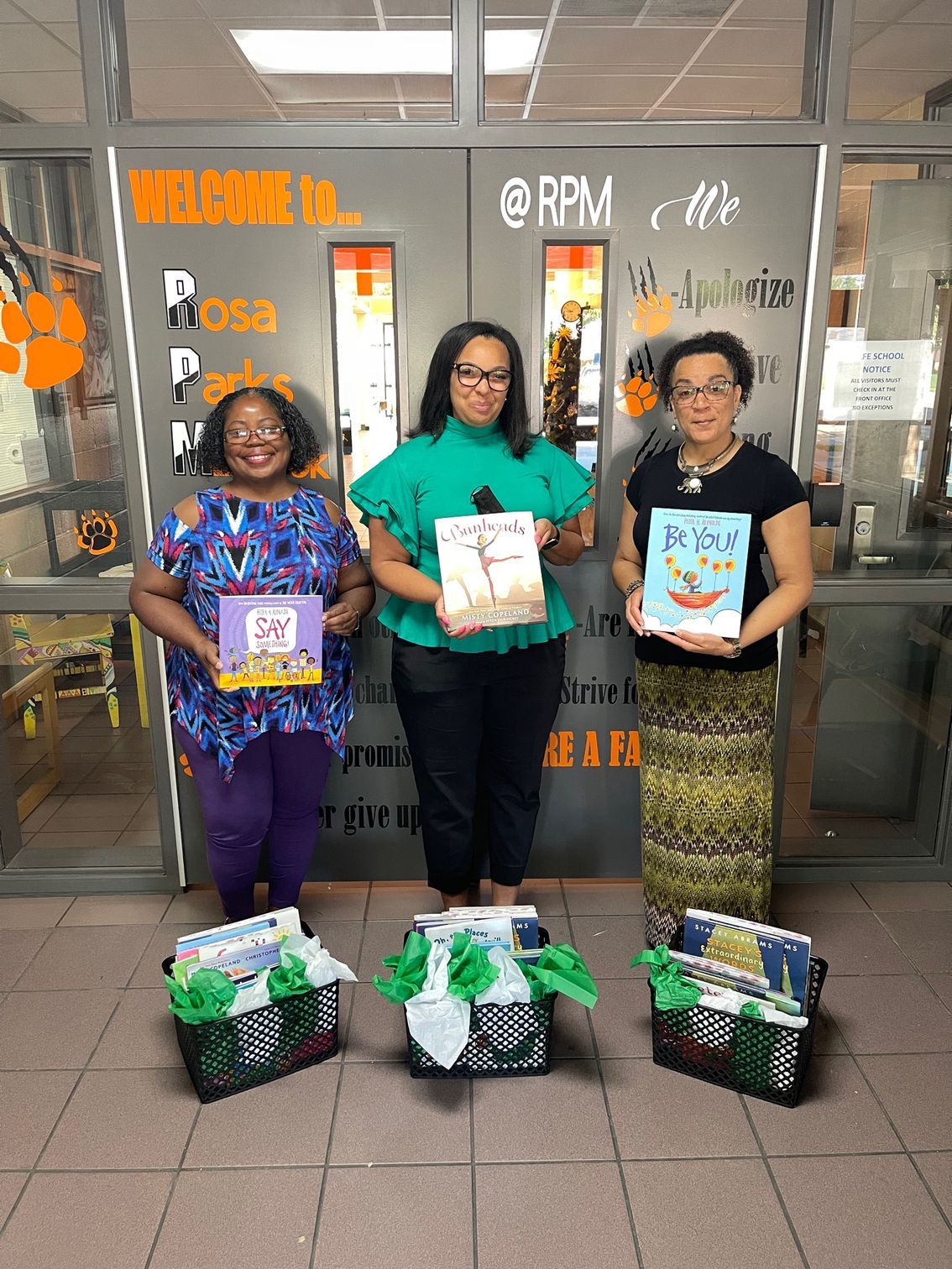 Three women holding books, standing by baskets of books in a school hallway with welcoming decorations.