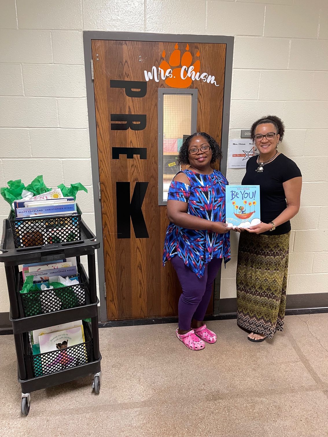 Two women stand by a Pre-K door, one holding a book. A cart with books and green decorations is nearby.