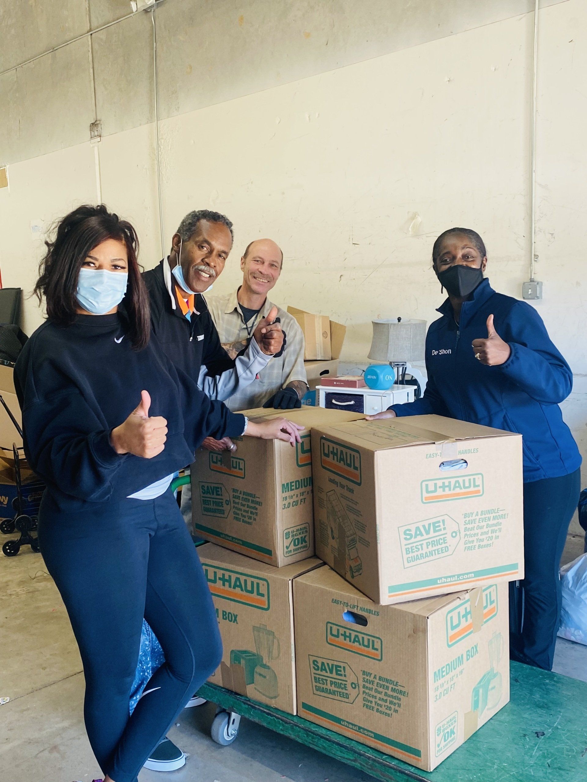 Four people pose with boxes stacked on a dolly, giving thumbs-up gestures. They are in a warehouse.