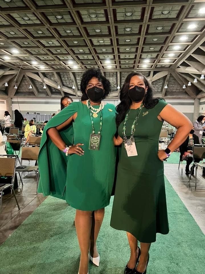 Two women in green dresses pose indoors, wearing masks, with event badges, and a green carpet.