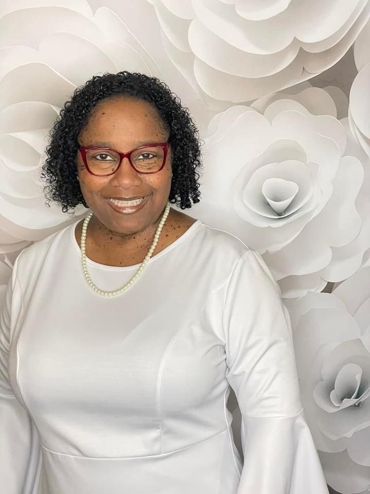 Woman in a white dress and red glasses smiles, wearing a pearl necklace, in front of white paper flowers.