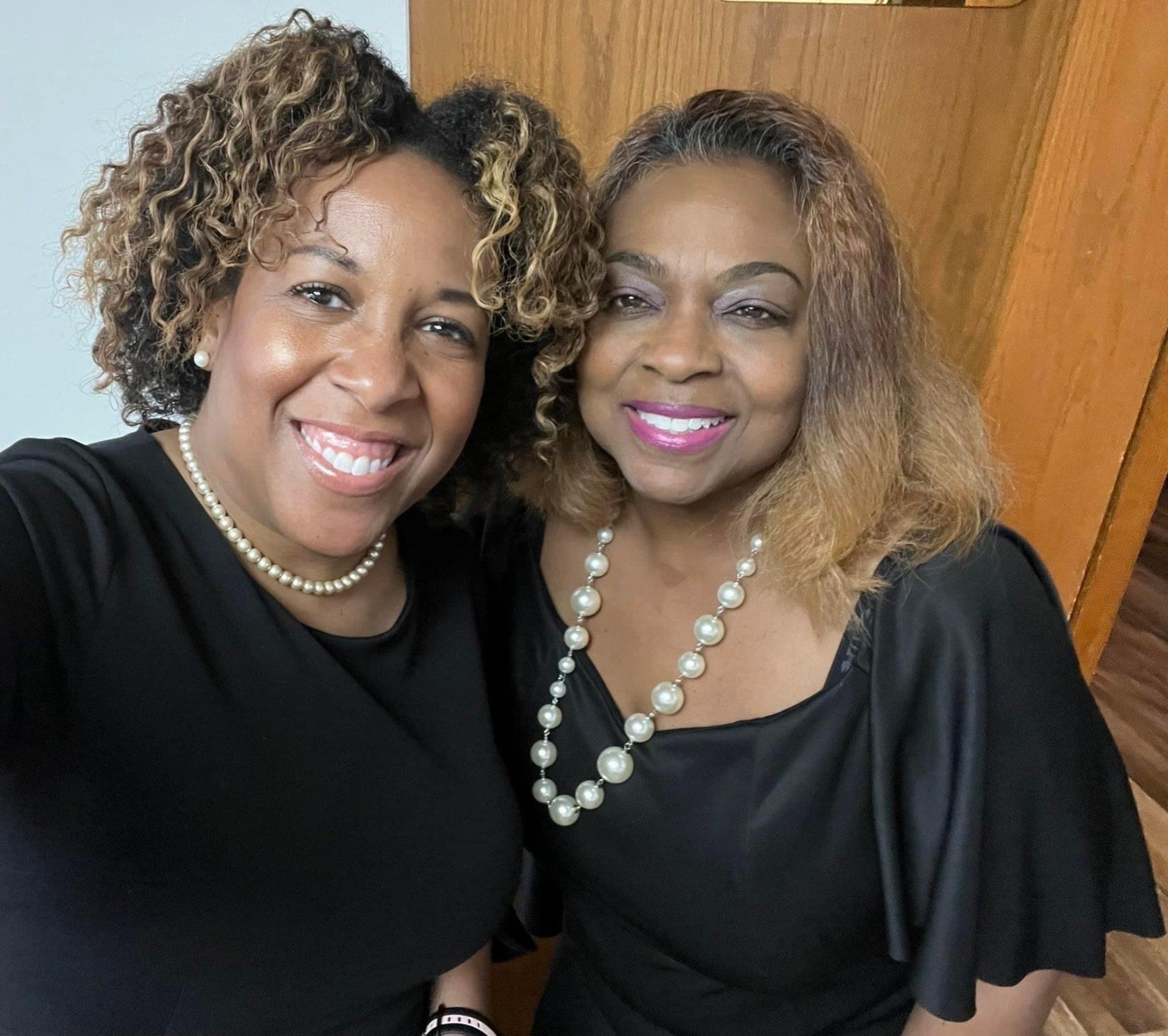 Two smiling women in black tops and pearl necklaces, indoors.