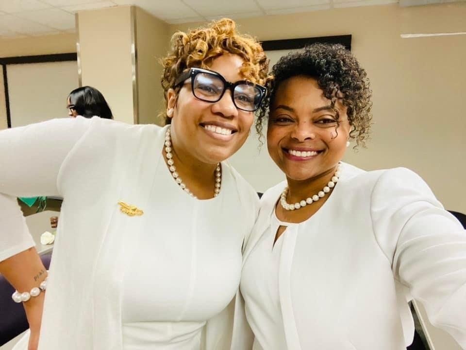 Two women in white outfits smile, indoors, one with glasses, both with pearl necklaces.
