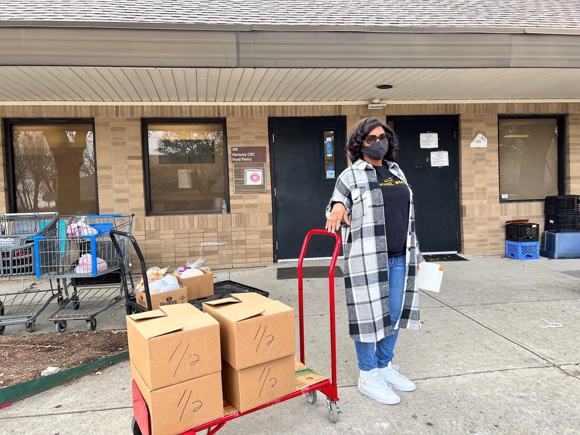 Woman with boxes on a cart outside a building, wearing a mask and holding papers.