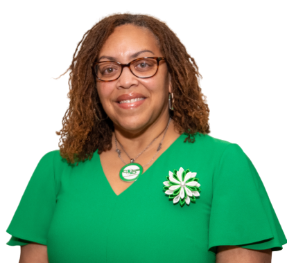Woman wearing glasses and a green dress, smiling, with a flower pin and necklace.