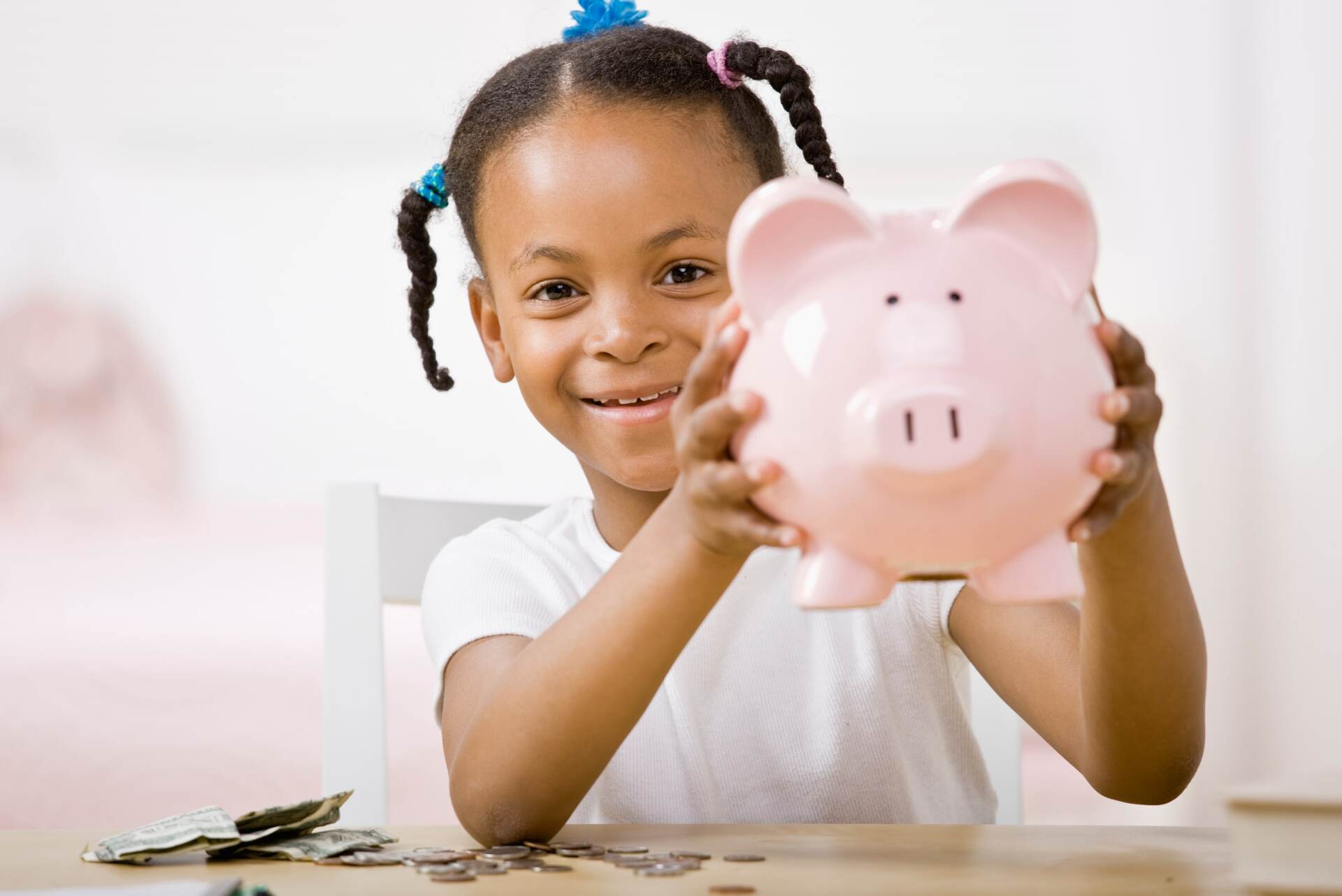 Girl holding a pink piggy bank, smiling, with coins on a table.