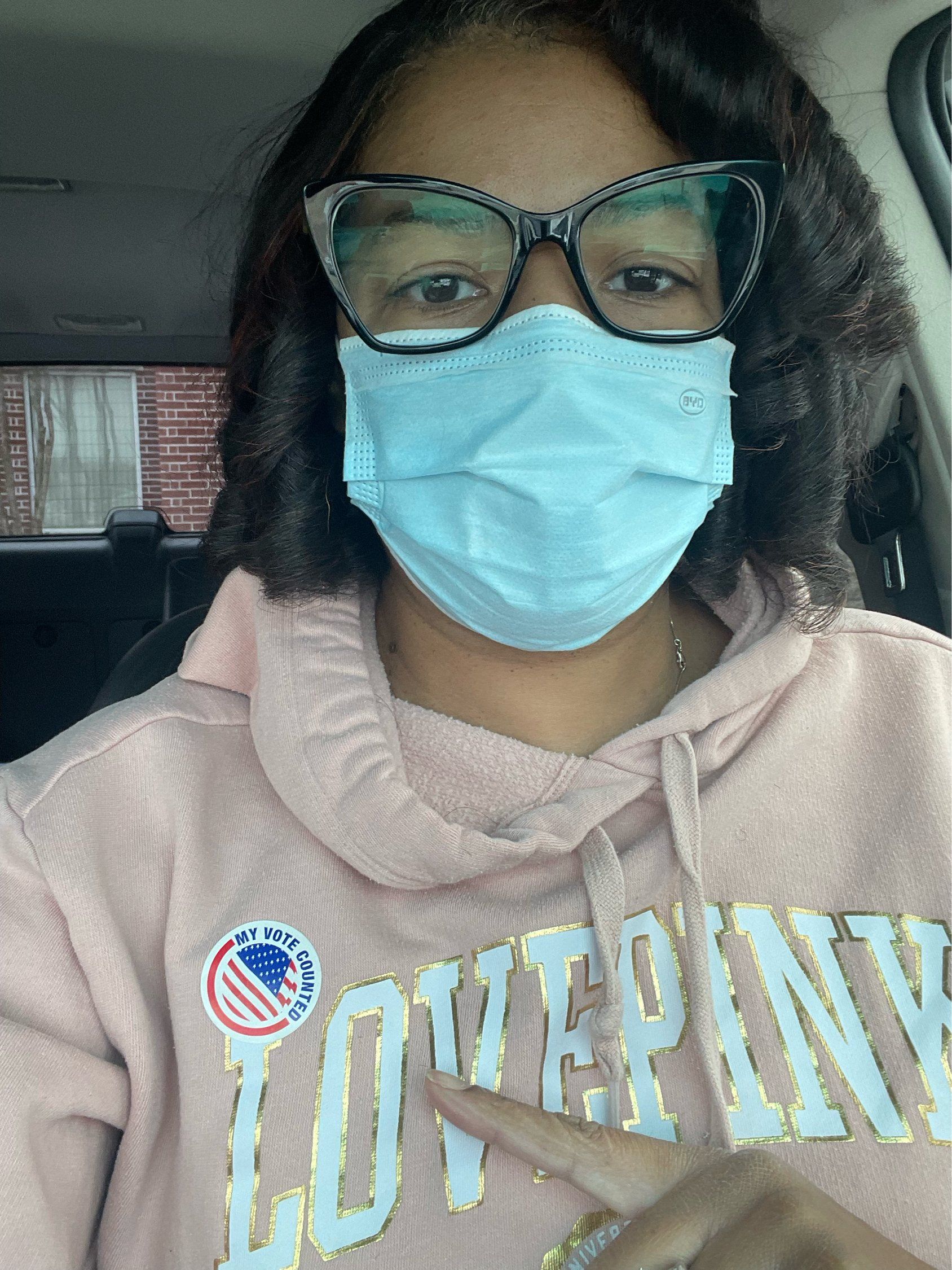 Woman in a pink hoodie wearing a mask and glasses, pointing at a 