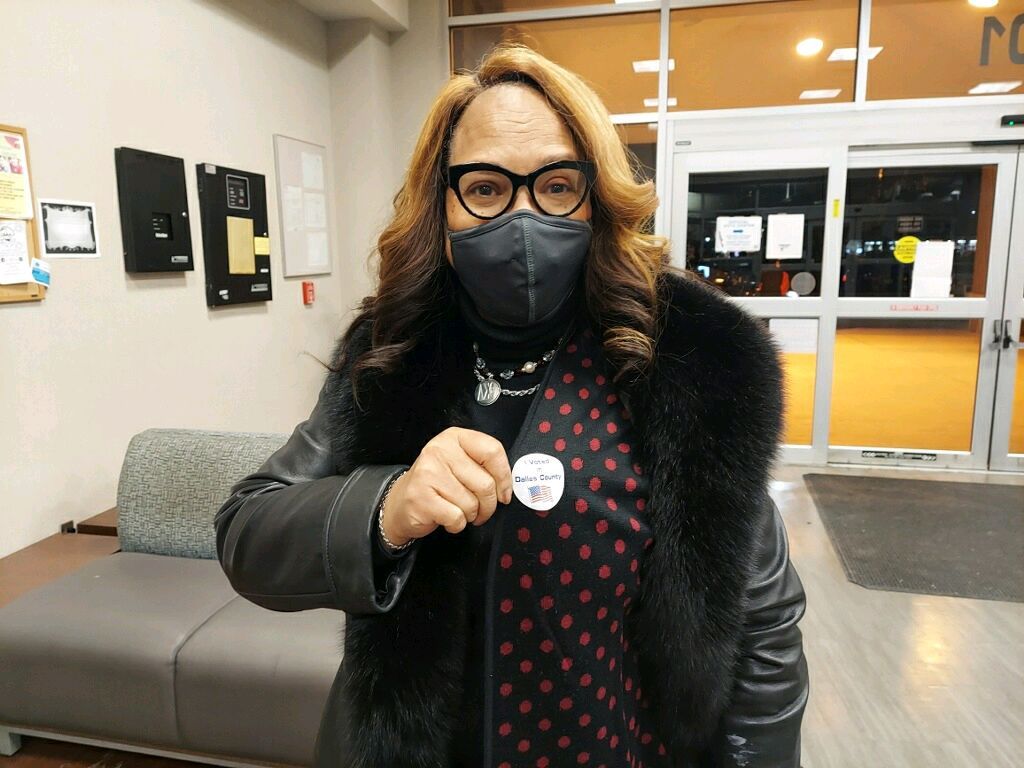 Person wearing a mask and fur-trimmed coat, holding a button. Indoors, near an entrance with glass doors.