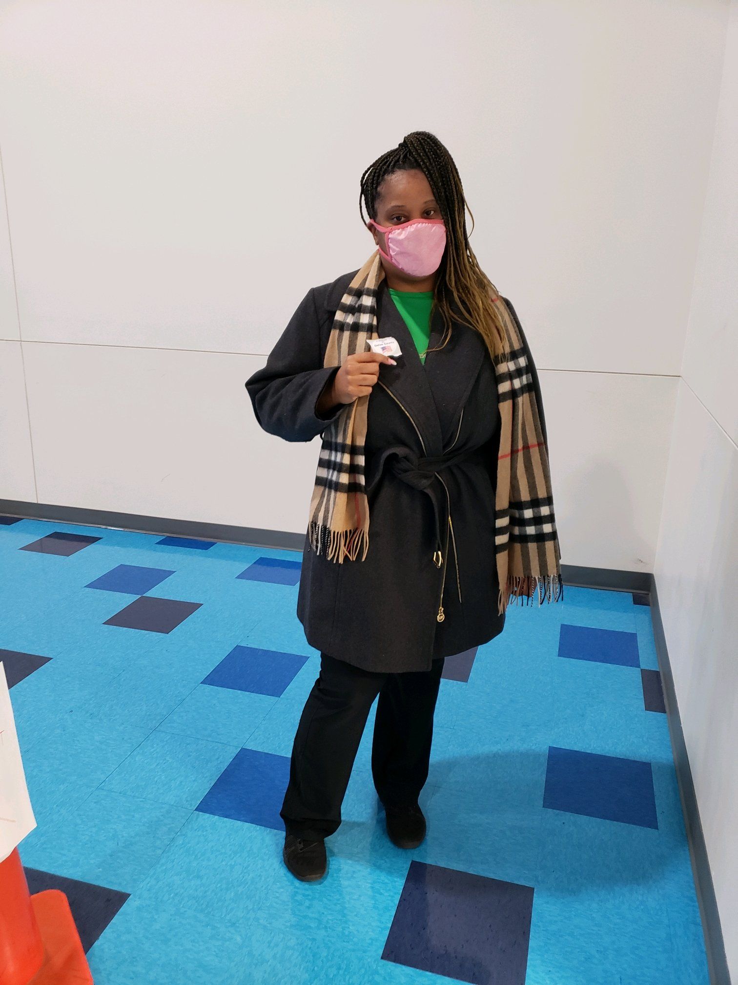 Woman in black coat and scarf wearing a pink face mask standing on blue and white flooring.