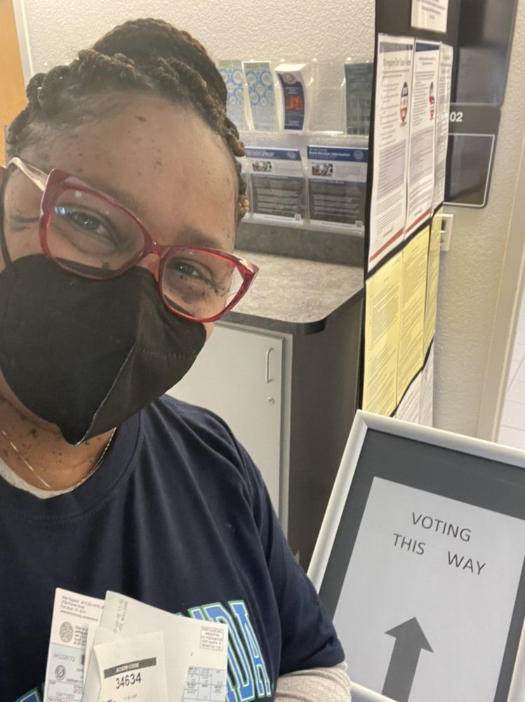 Person wearing a mask and glasses at a voting location, holding ballots, next to a 