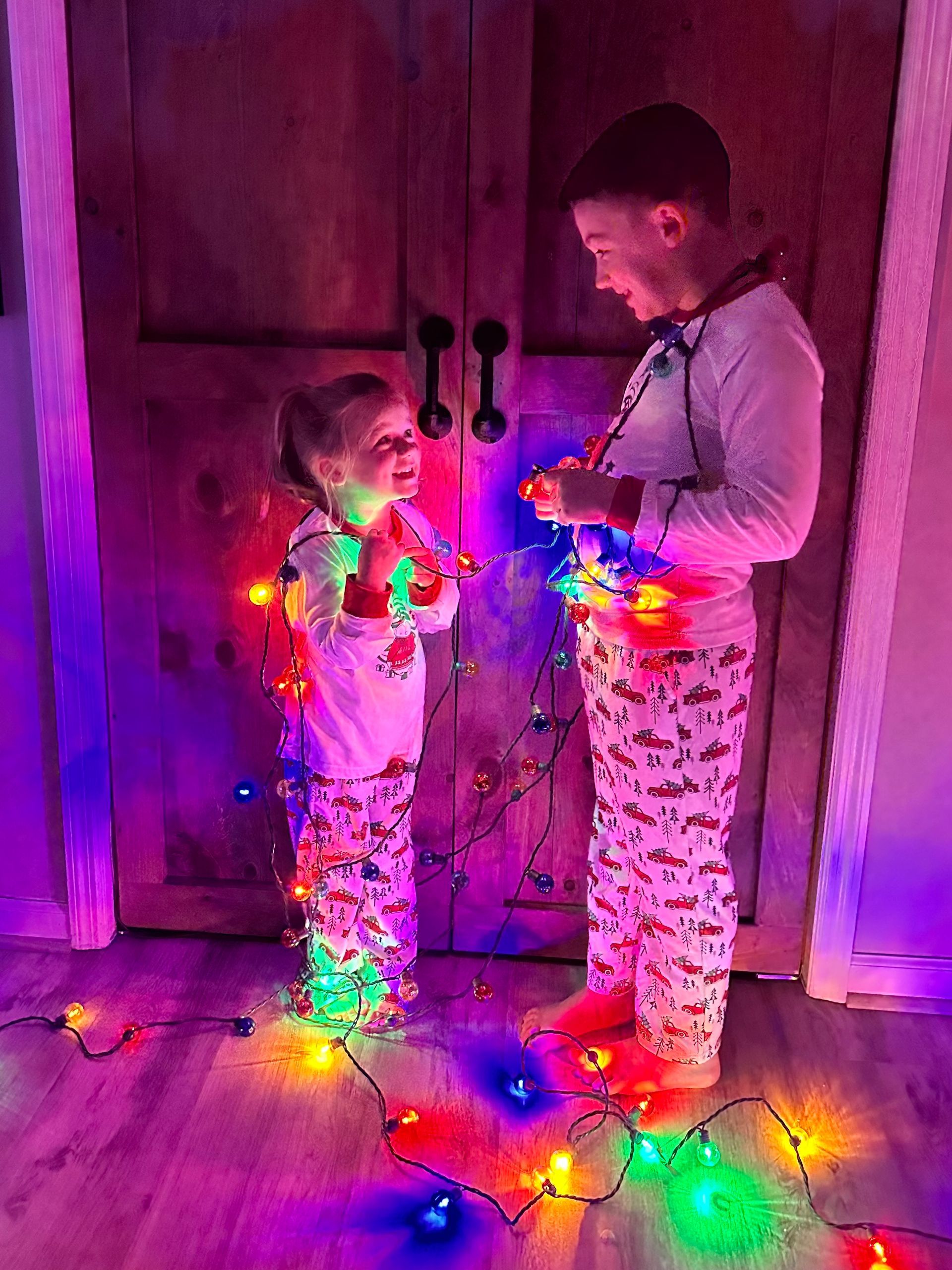 A boy and a girl are playing with christmas lights in a room.