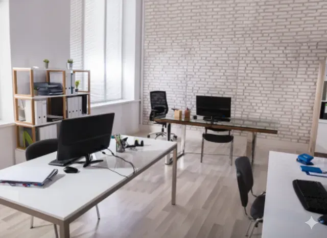 Office interior with desks, computers, and storage against a white brick wall and window.