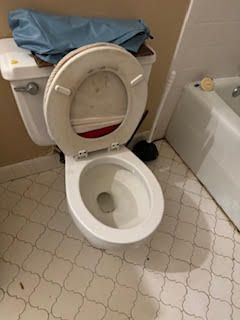 Toilet with seat up, partially open, in a bathroom with patterned floor and a bathtub visible in the background.