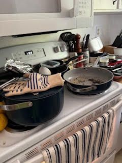 Cluttered kitchen stovetop with dirty pan, pot, utensils, and oven mitt; a messy cooking scene.