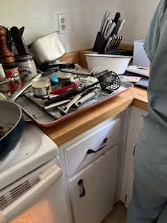 Cluttered kitchen counter with utensils, a tray of items, and a white stove. A person in the right.