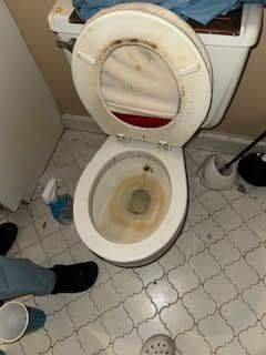 Dirty toilet with stained bowl and seat, bathroom floor, cleaning spray and foot in frame.