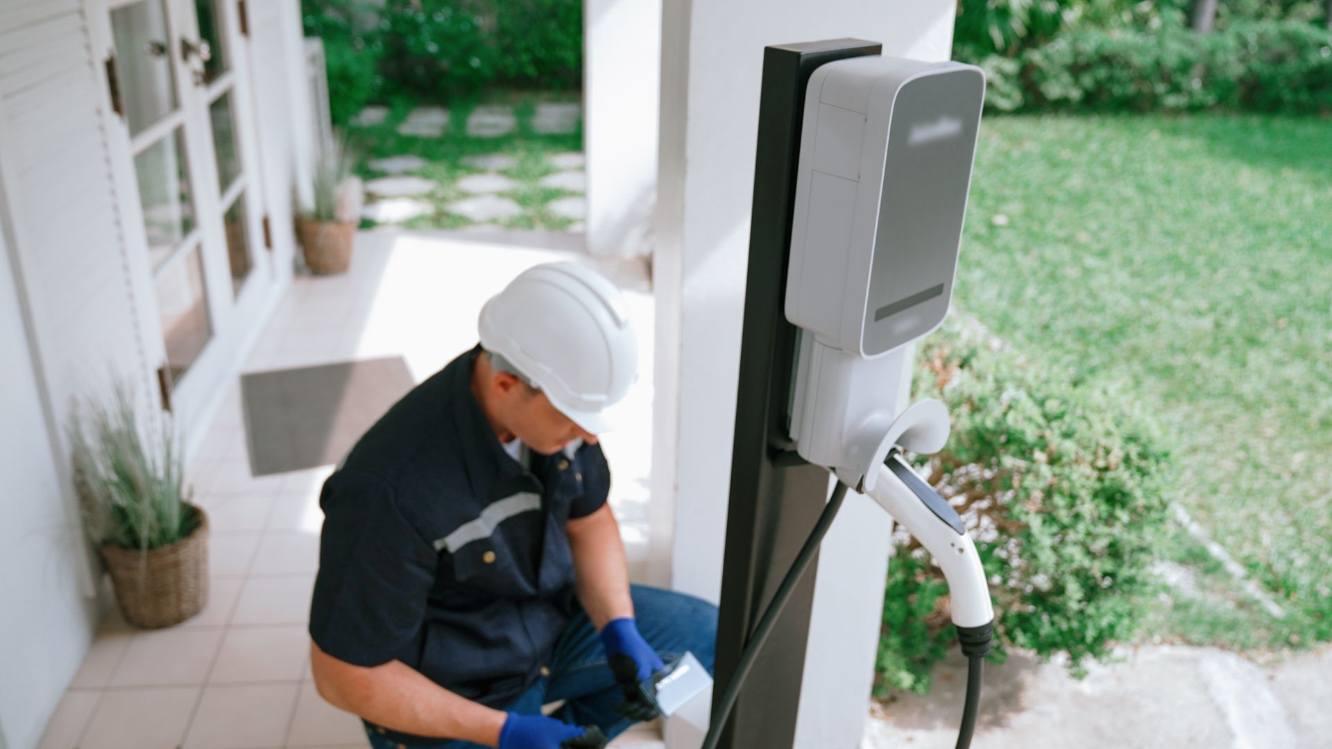 Technician in hard hat installing an outdoor EV charger beside a porch